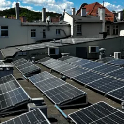 Instalacja fotowoltaiczna na dachu budynku z widocznymi panelami słonecznymi, klimatyzatorami i budynkami w tle, w słoneczny dzień.