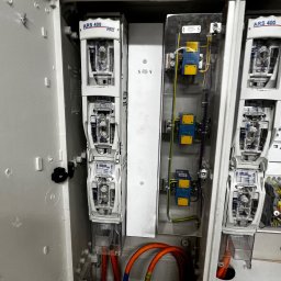 CENTRUM REALIZACJI PROSTA SPÓŁKA AKCYJNA - Otwarta szafa elektryczna z bezpiecznikami ARS 400 PRO, widoczne kable w kolorze pomarańczowym i niebieskie zabezpieczenia. Instalacja wewnątrz białej obudowy.