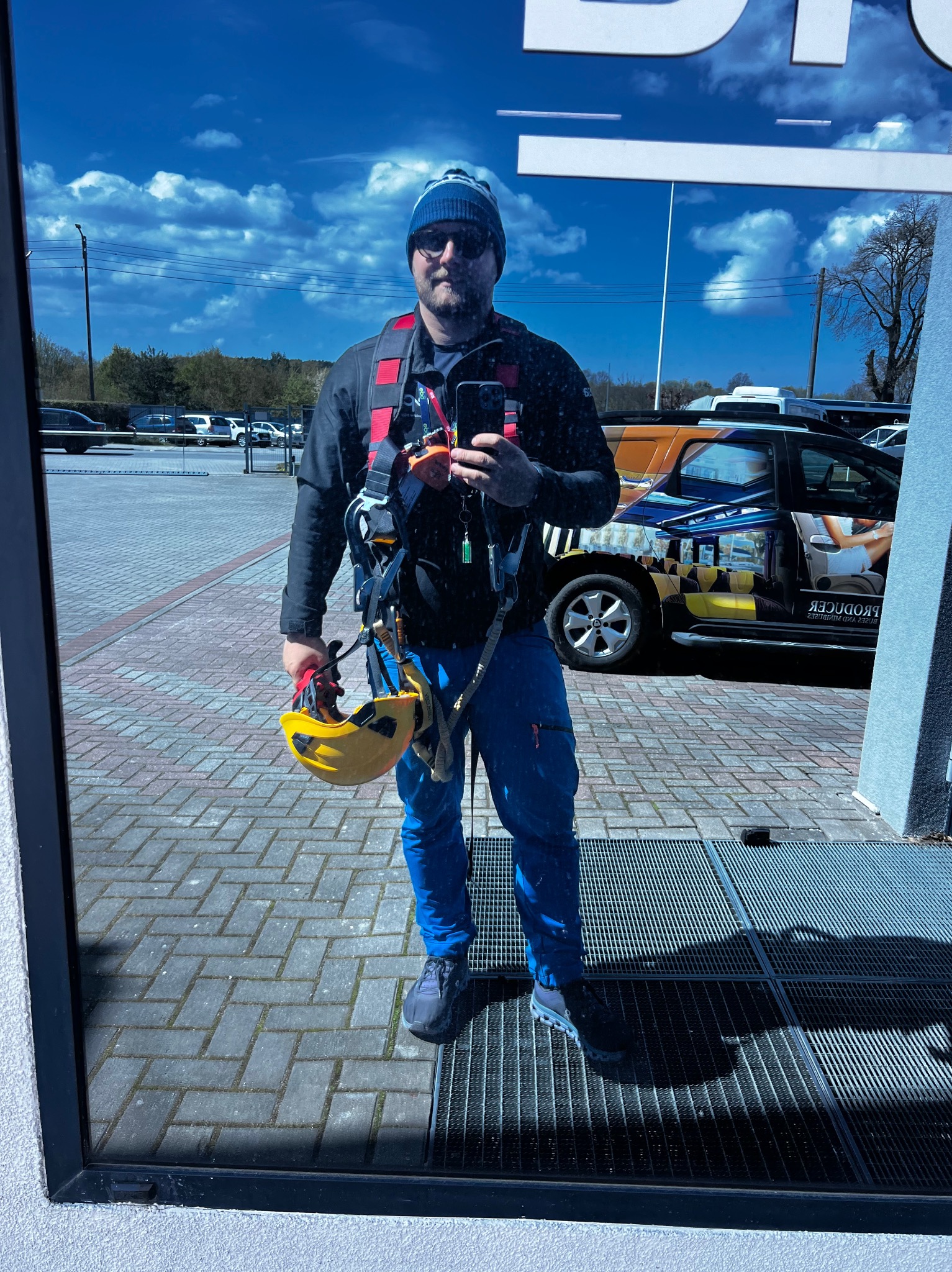 Autoportret instalatora w uprzęży i kasku, odbicie w szybie na tle parkingu i błękitnego nieba z chmurami. Widoczny samochód z grafiką.