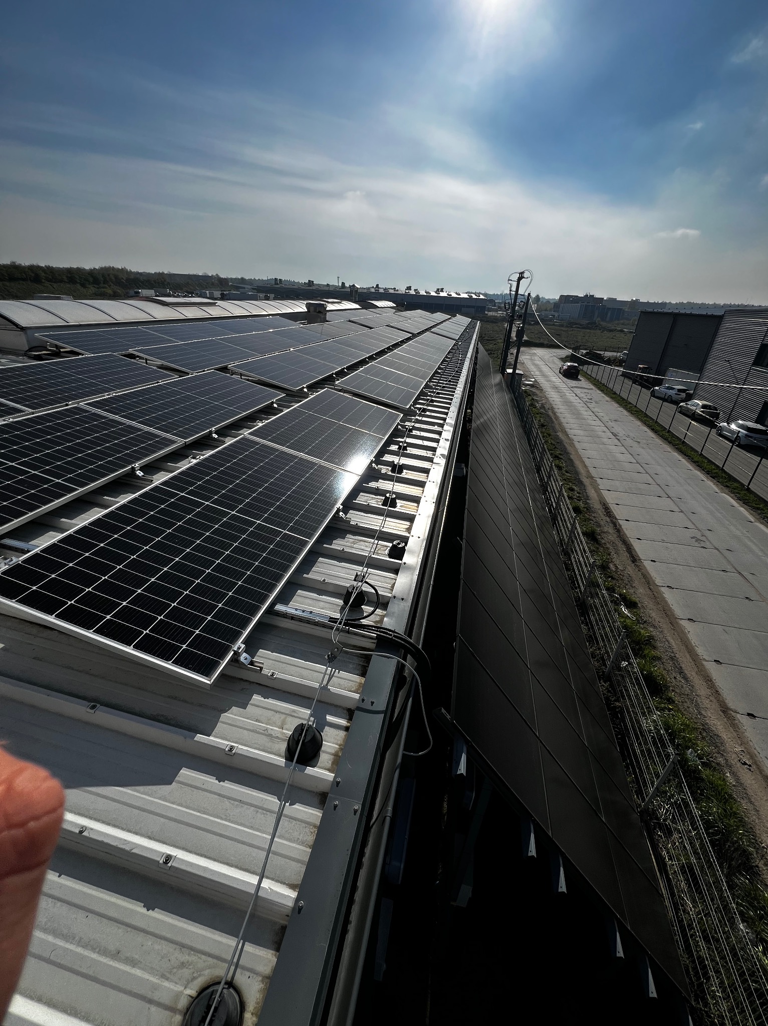 Rząd paneli słonecznych na dachu budynku przemysłowego w słoneczny dzień. Widoczny fragment dłoni w lewym dolnym rogu. Perspektywa z góry.