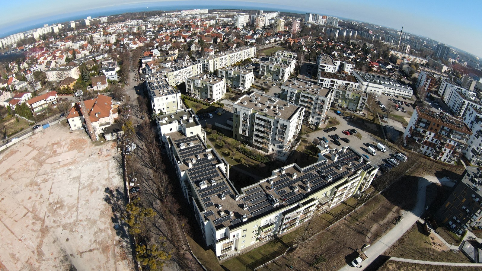 Widok z drona na osiedle z panelami słonecznymi na dachach budynków. W tle zabudowa miejska i linia horyzontu z morzem. Słoneczny dzień, perspektywa z góry.