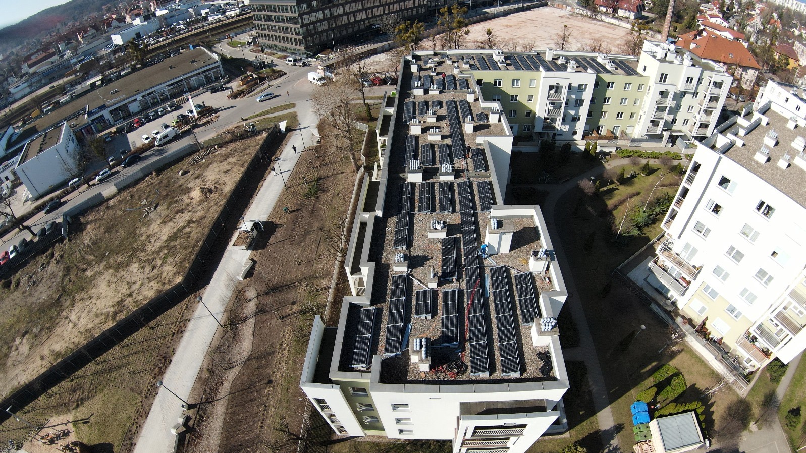 Widok z góry na panele słoneczne na dachu budynku mieszkalnego, z widocznymi elementami instalacji i otaczającą zabudową miejską w słoneczny dzień.