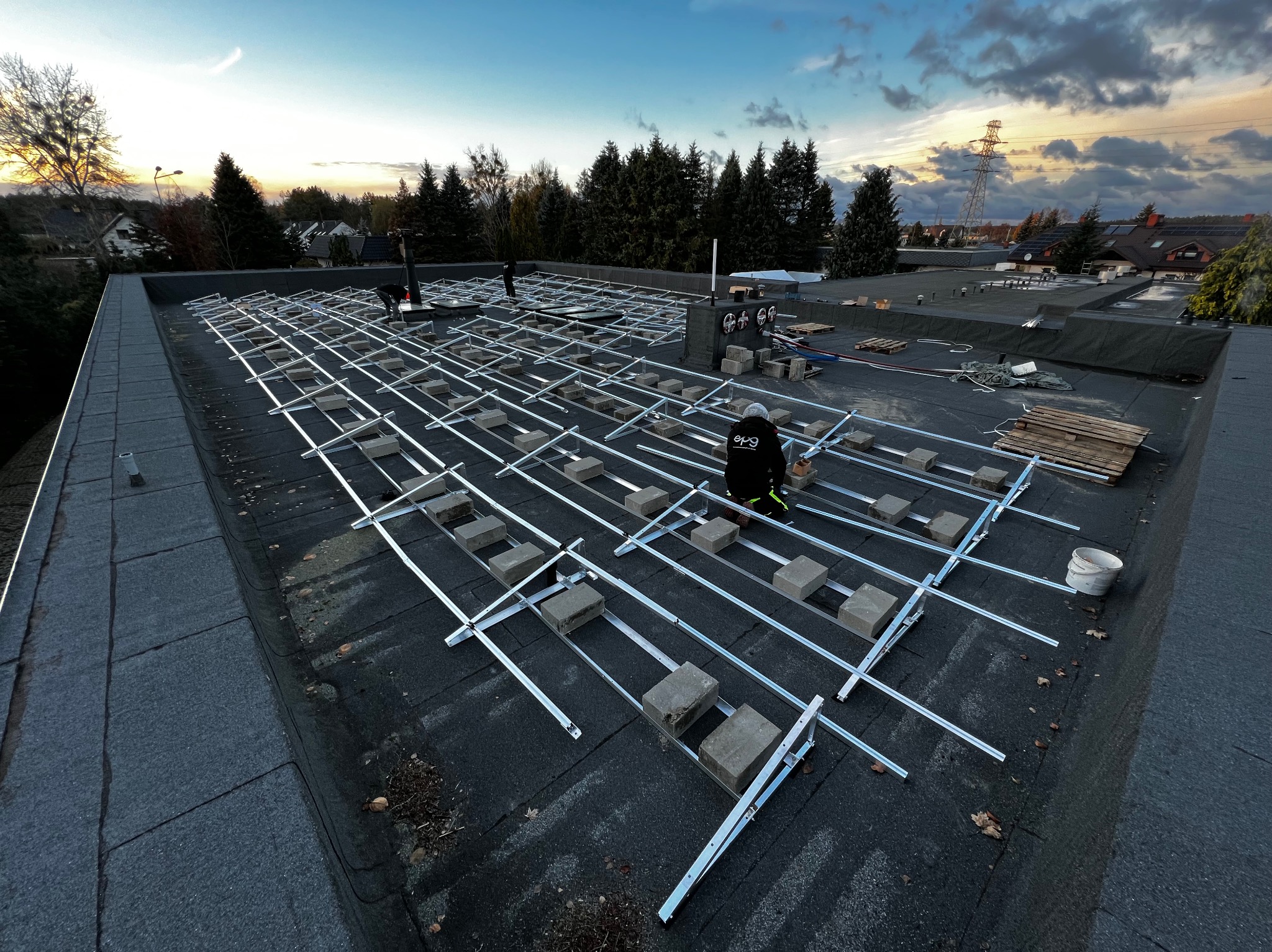 Montaż konstrukcji wsporczej pod panele słoneczne na dachu budynku. Widoczne metalowe profile, betonowe bloczki i pracownik w stroju roboczym. Tło: drzewa i zachodzące słońce.