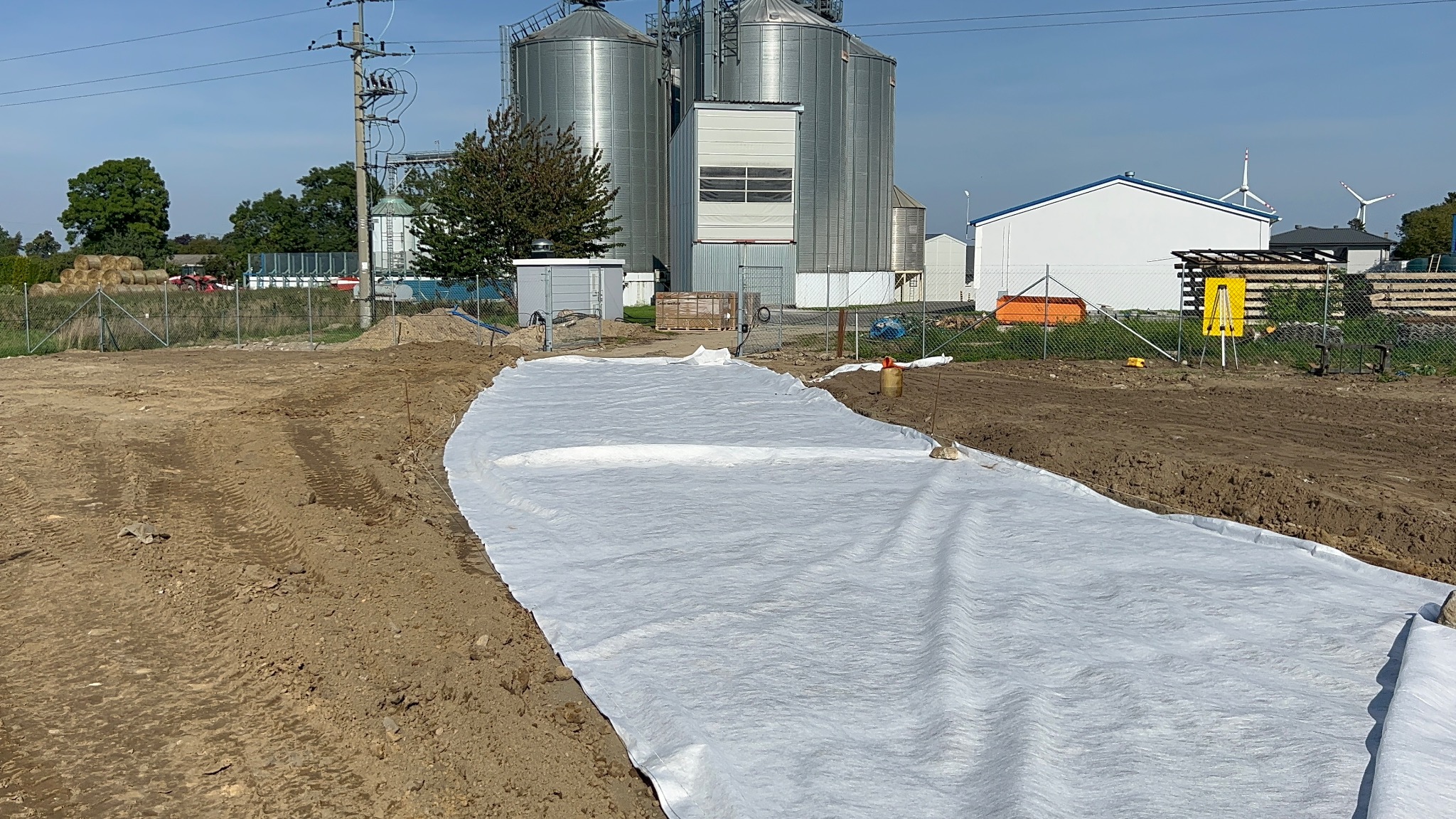 Przygotowanie terenu pod instalację: rozłożona biała geowłóknina na wykopie, w tle silosy zbożowe i budynek z turbinami wiatrowymi.