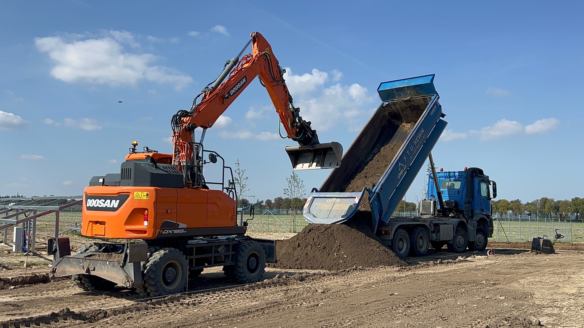 Pomarańczowa koparka ładuje ziemię do niebieskiej wywrotki na placu budowy z widocznymi w tle panelami fotowoltaicznymi w trakcie montażu.