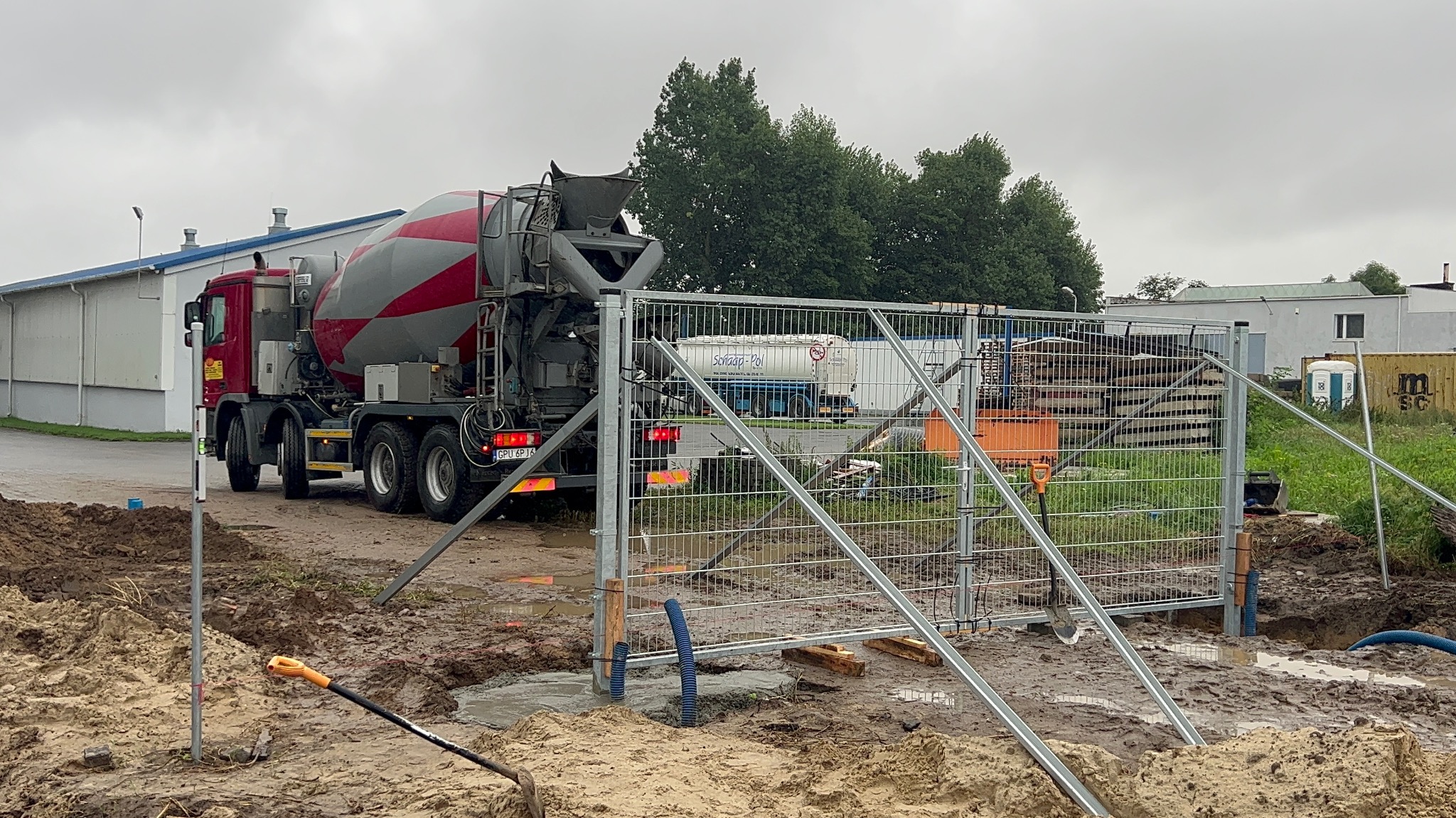 Betoniarka w trakcie wylewania fundamentów pod ogrodzenie na placu budowy, widoczne narzędzia i tymczasowe zabezpieczenia.