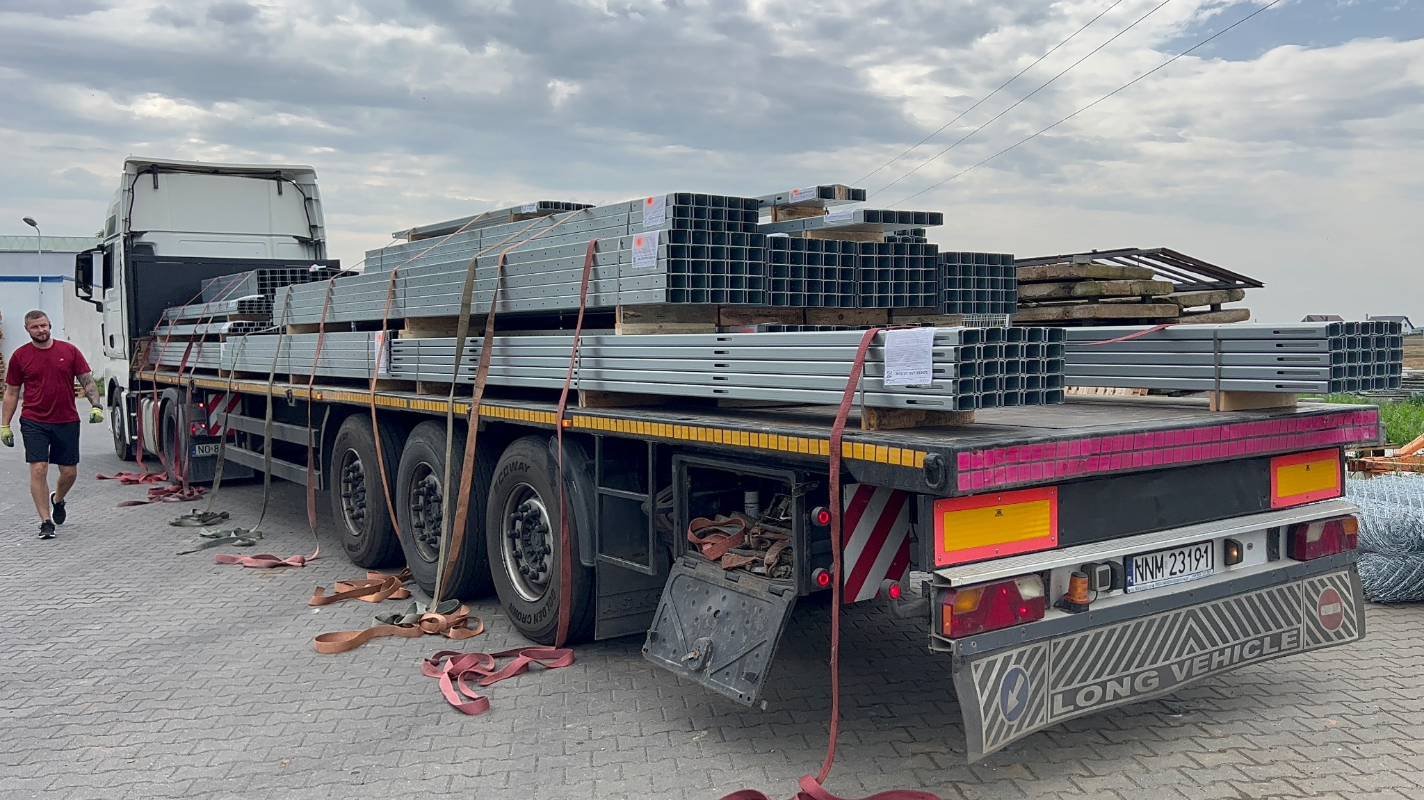 Transport profili konstrukcyjnych na naczepie ciężarówki, zabezpieczonych pasami transportowymi. Widoczny fragment tablicy rejestracyjnej i sylwetka mężczyzny.