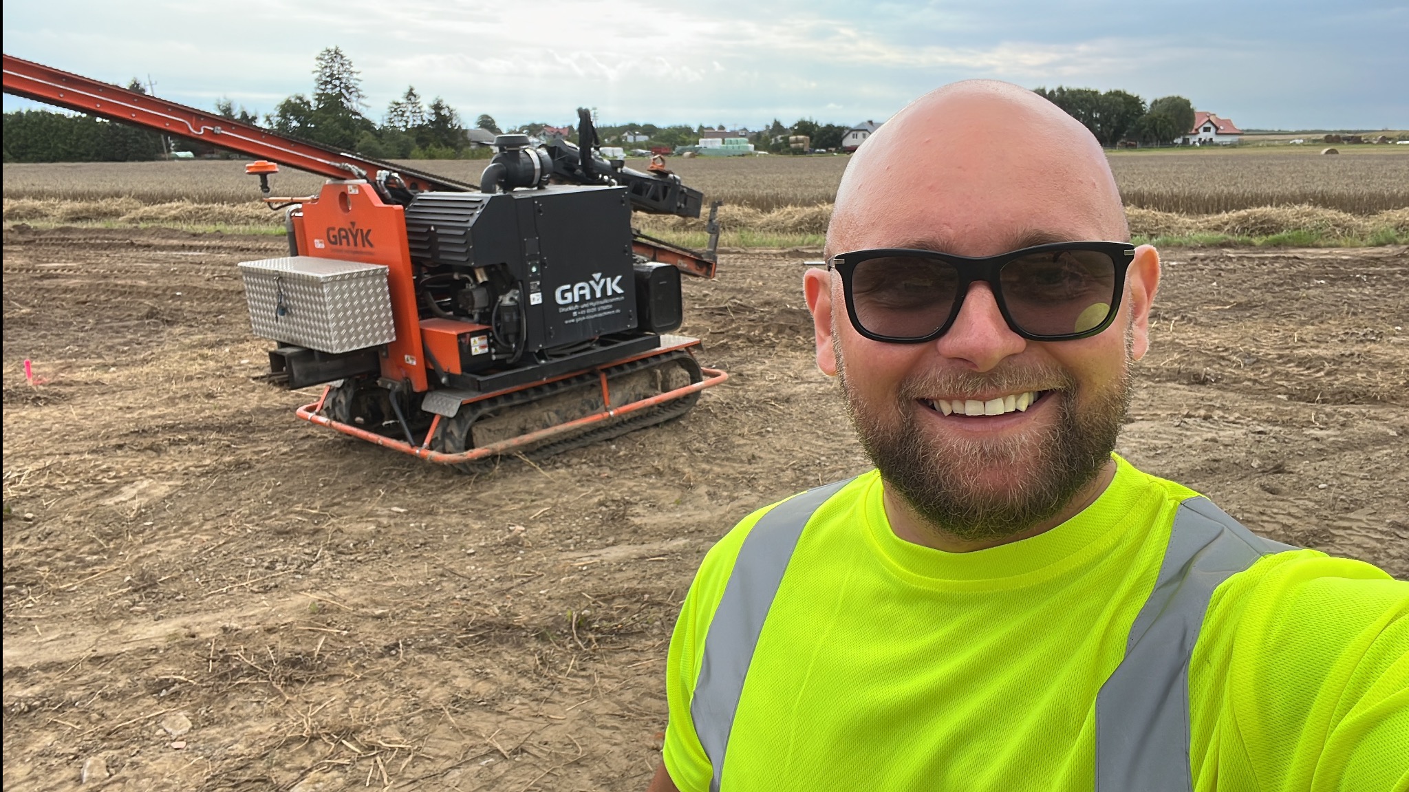 Mężczyzna w okularach i jaskrawej koszulce pozuje na tle pomarańczowej wiertnicy geologicznej na gąsienicach, stojącej na polu z niską roślinnością.