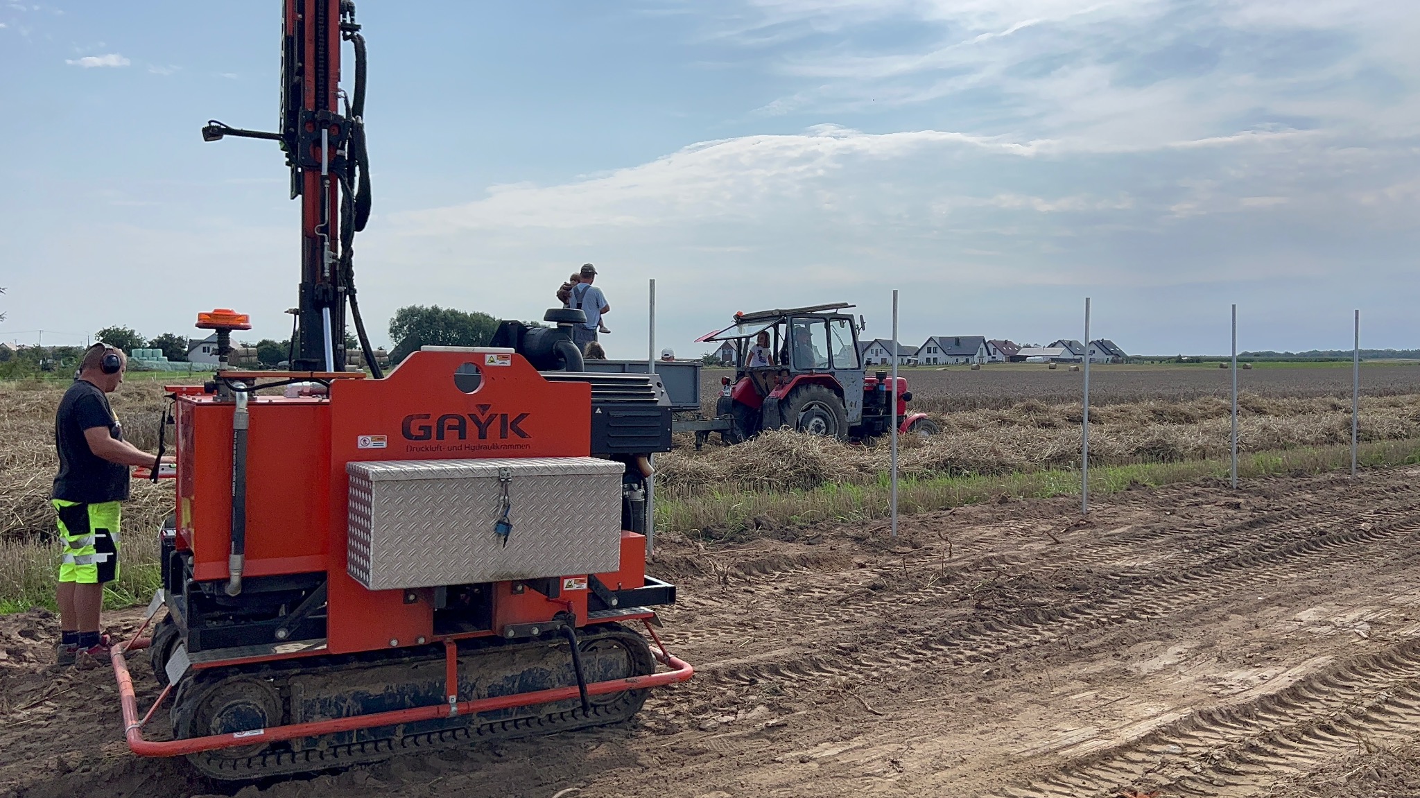 Montaż słupków w polu z użyciem specjalistycznej maszyny GAYK i traktora. Widoczne ustawione słupki, pole po żniwach i domy w oddali. Praca w toku.