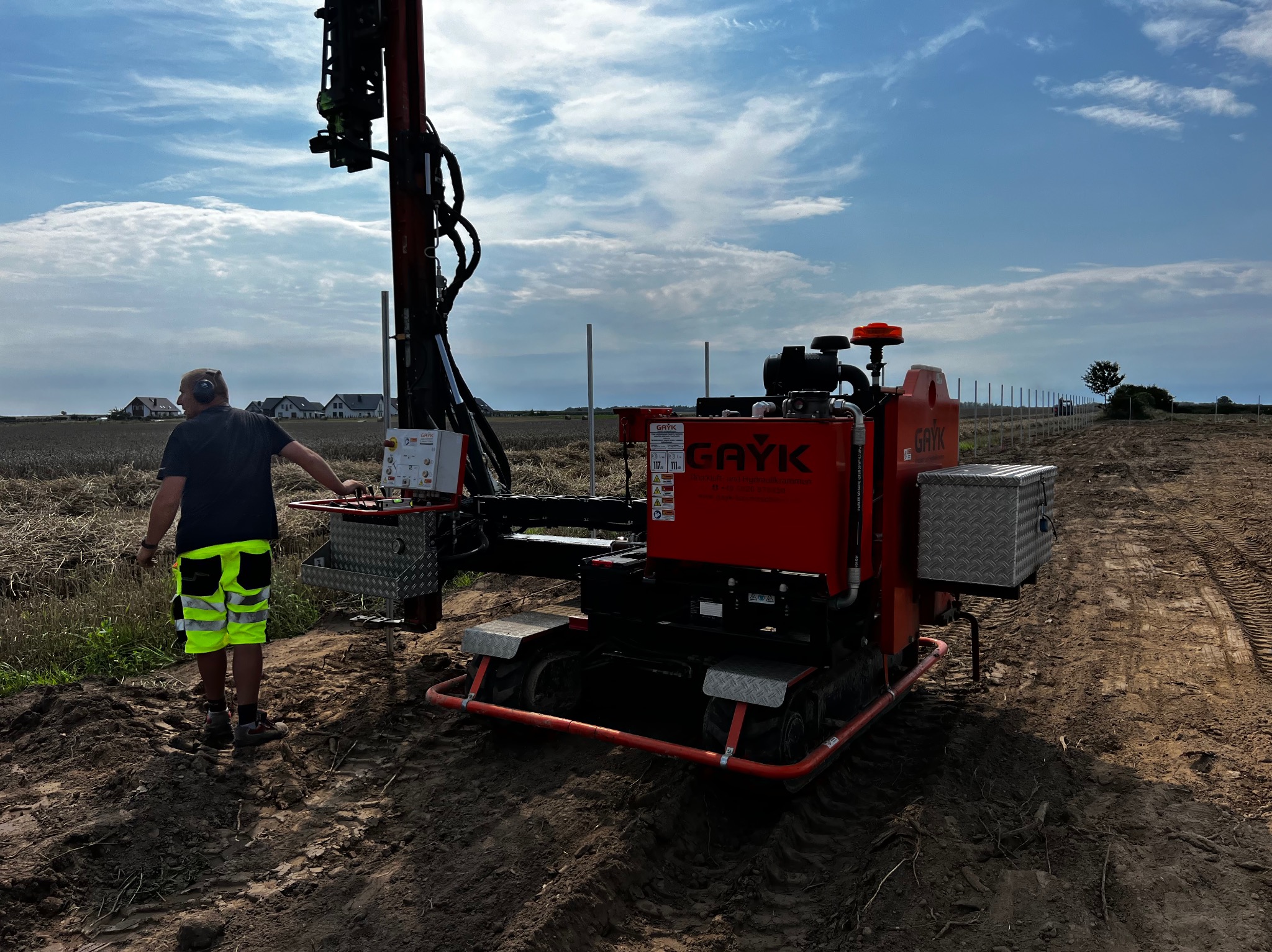 Pracownik obsługuje czerwoną maszynę GAYK do wbijania słupków pod panele fotowoltaiczne na polu, widoczne domy w tle, błękitne niebo.