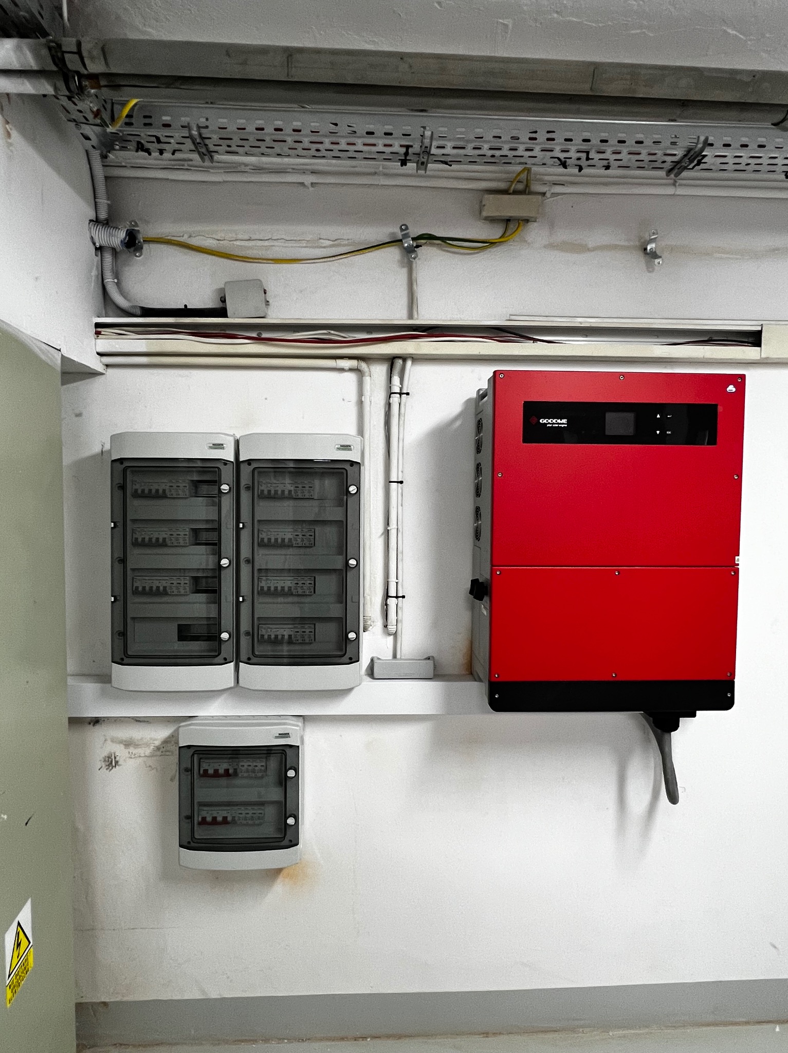 Czerwony inwerter GoodWe, dwie szare skrzynki elektryczne i mniejsza skrzynka na ścianie w pomieszczeniu. Widoczne kable i metalowe korytka kablowe.