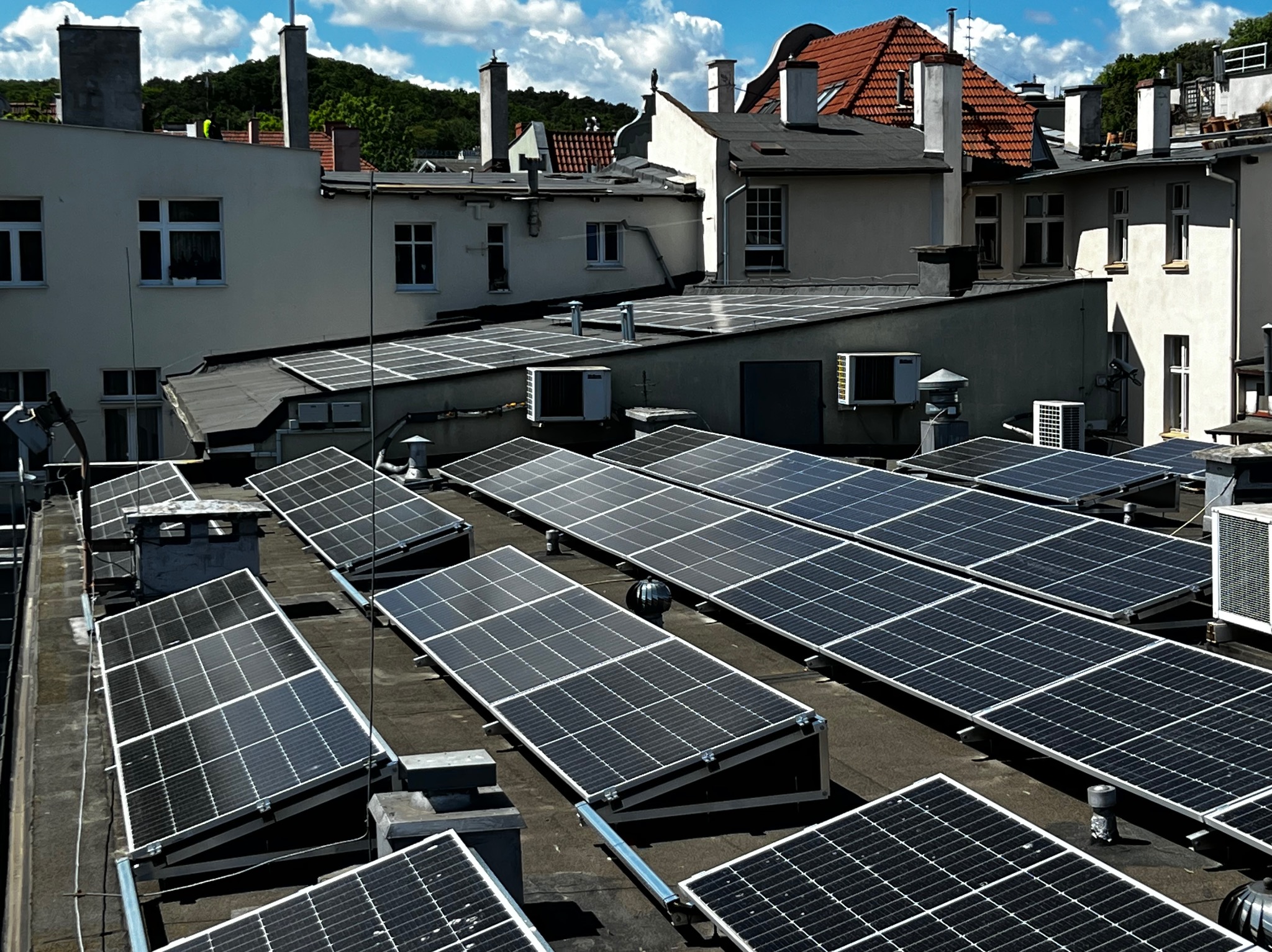 Instalacja fotowoltaiczna na dachu budynku z widocznymi panelami słonecznymi, klimatyzatorami i budynkami w tle, w słoneczny dzień.
