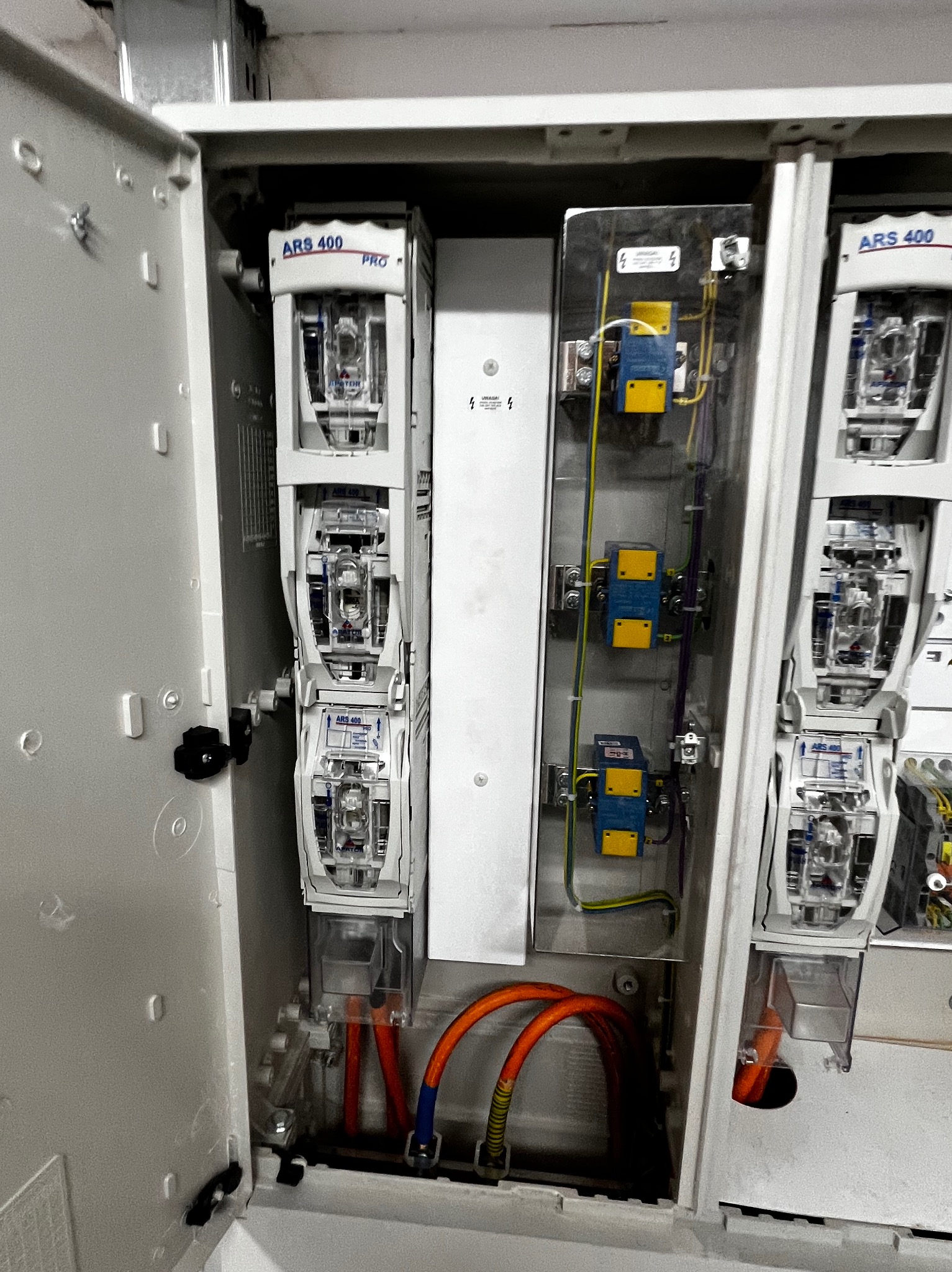 Otwarta szafa elektryczna z bezpiecznikami ARS 400 PRO, widoczne kable w kolorze pomarańczowym i niebieskie zabezpieczenia. Instalacja wewnątrz białej obudowy.