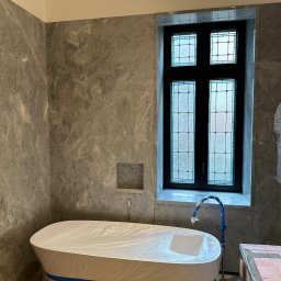 Perfection pro design ING - Wykończona łazienka w szarościach z wanną zabezpieczoną folią i oknem ze szprosami. W tle ściana wyłożona płytami imitującymi marmur.
