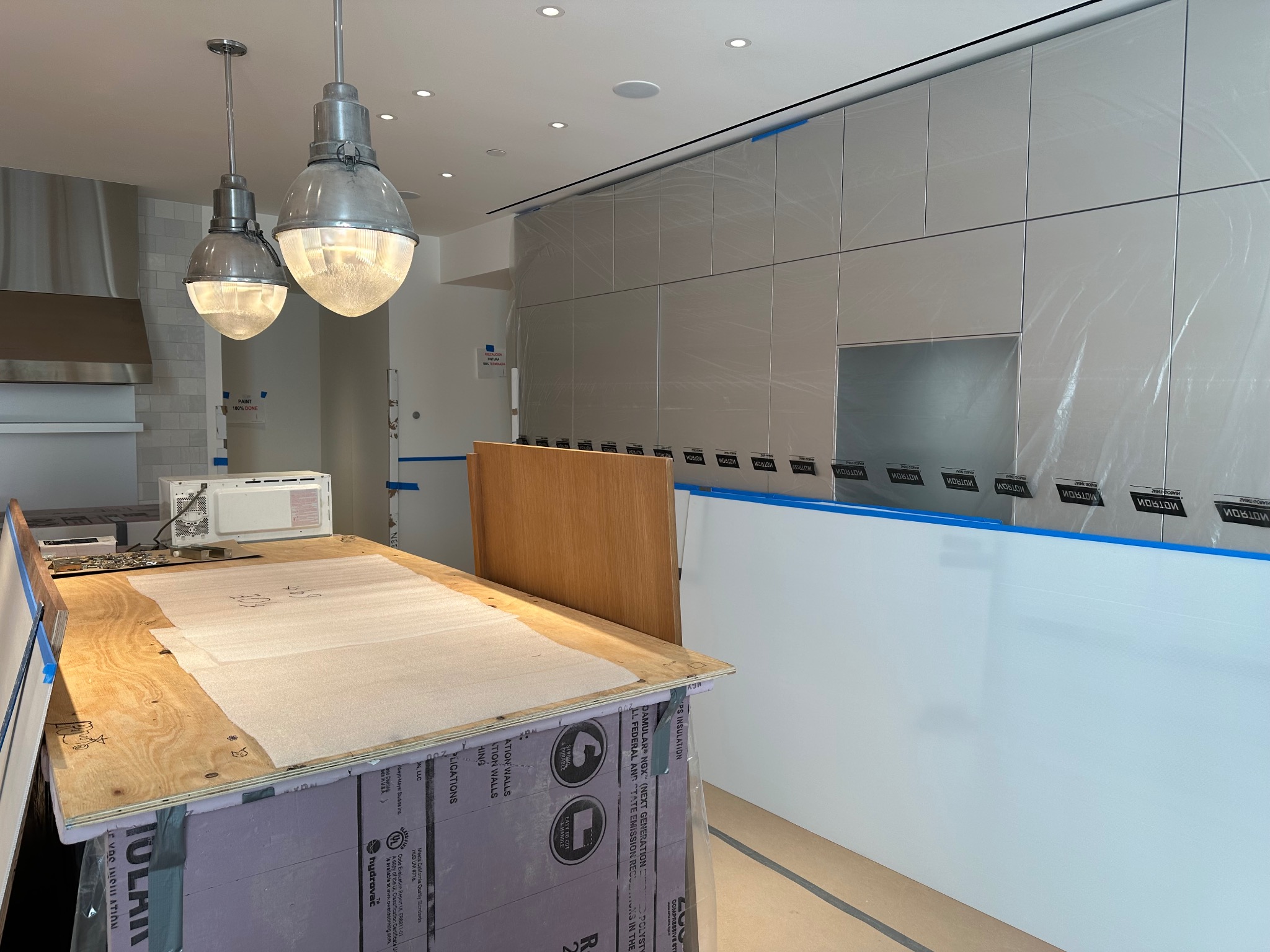 Wnętrze kuchni w trakcie prac wykończeniowych: zabezpieczone szafki, roboczy blat z mikrofalówką, wiszące lampy w stylu industrialnym. Widoczne materiały budowlane.