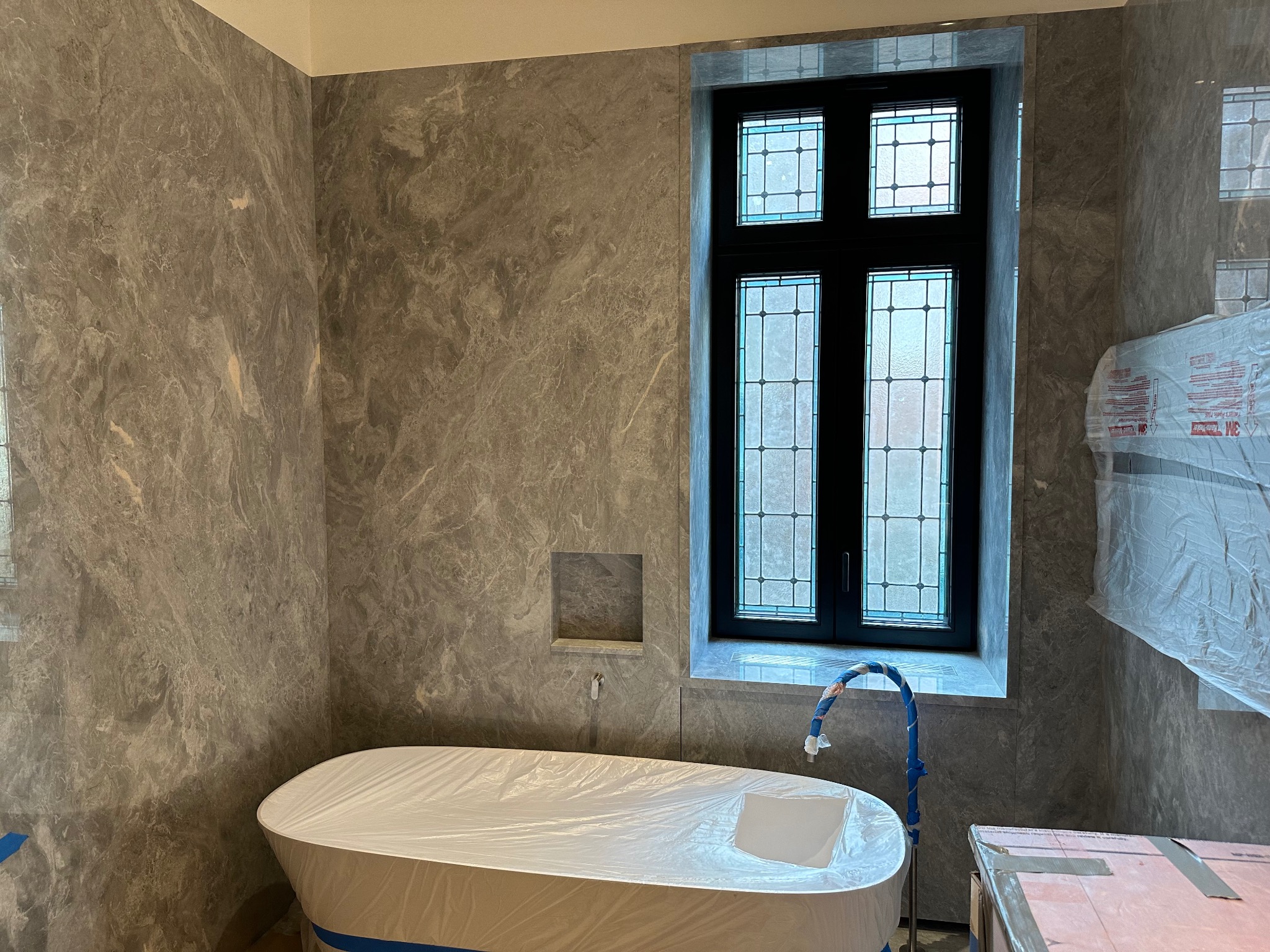 Wykończona łazienka w szarościach z wanną zabezpieczoną folią i oknem ze szprosami. W tle ściana wyłożona płytami imitującymi marmur.