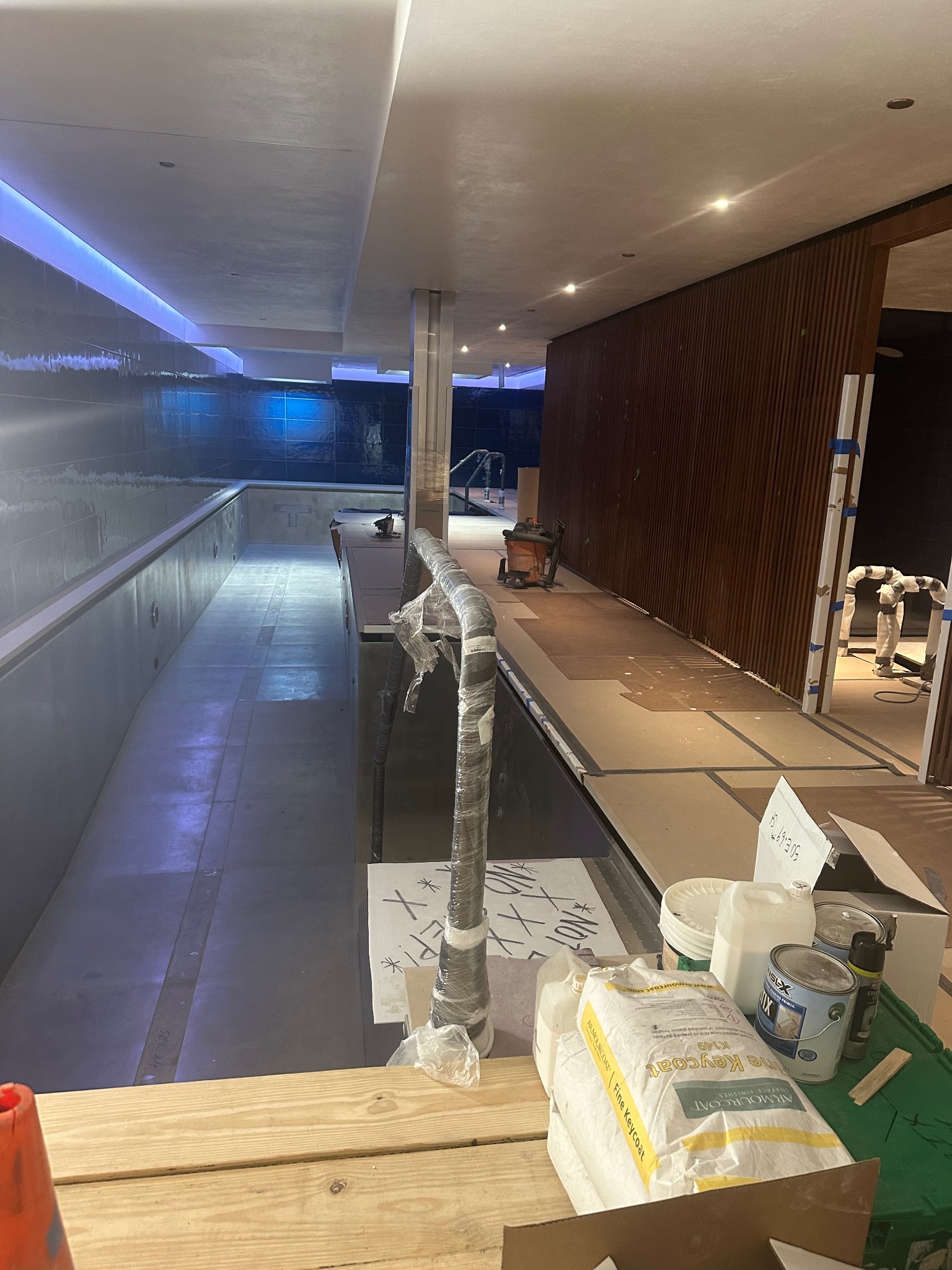 Wnętrze w trakcie wykończenia basenu z niebieskim oświetleniem, widoczne materiały budowlane, narzędzia i pracownicy w kombinezonach ochronnych.
