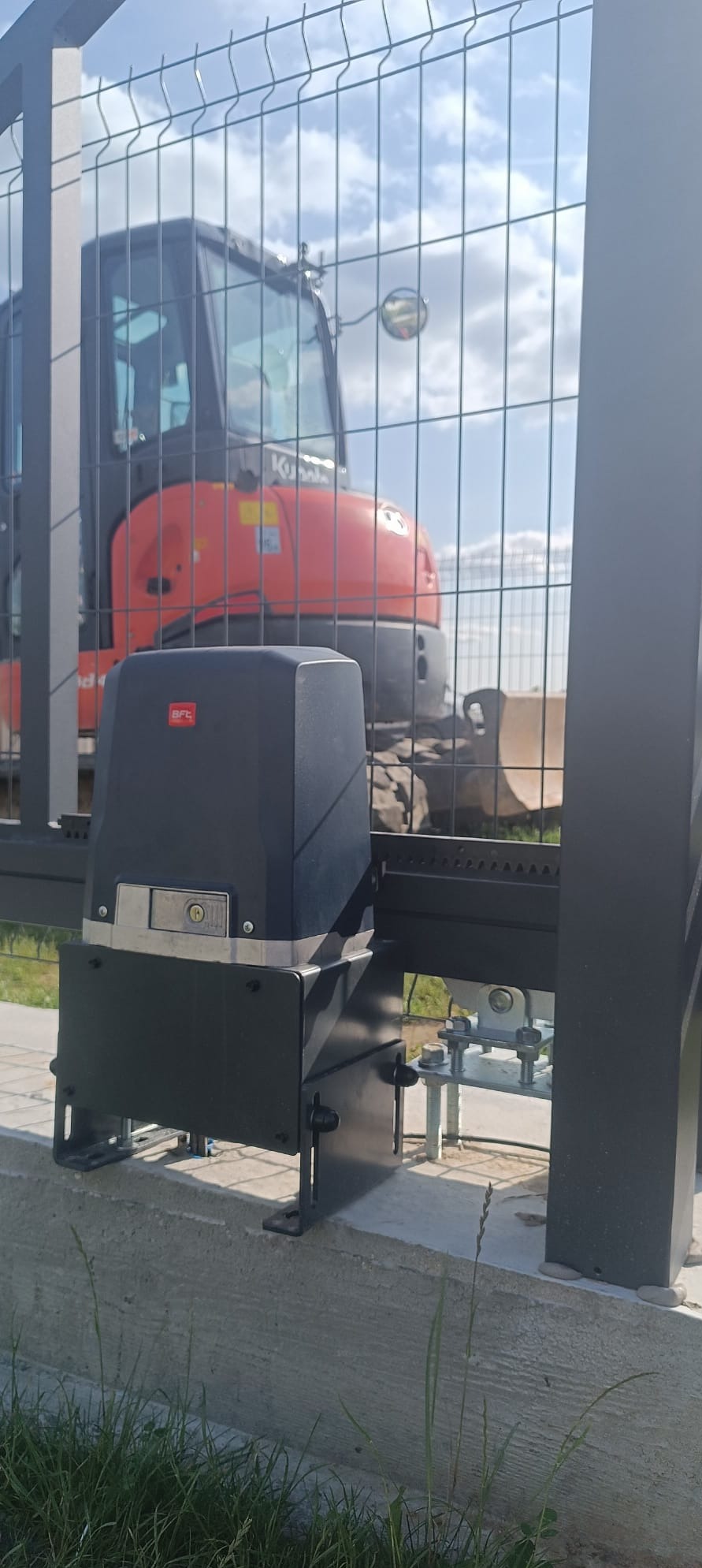 Zamontowany napęd bramy BFT przy ogrodzeniu panelowym, z widoczną koparką Kubota w tle i błękitnym niebem. Automatyka do bram.