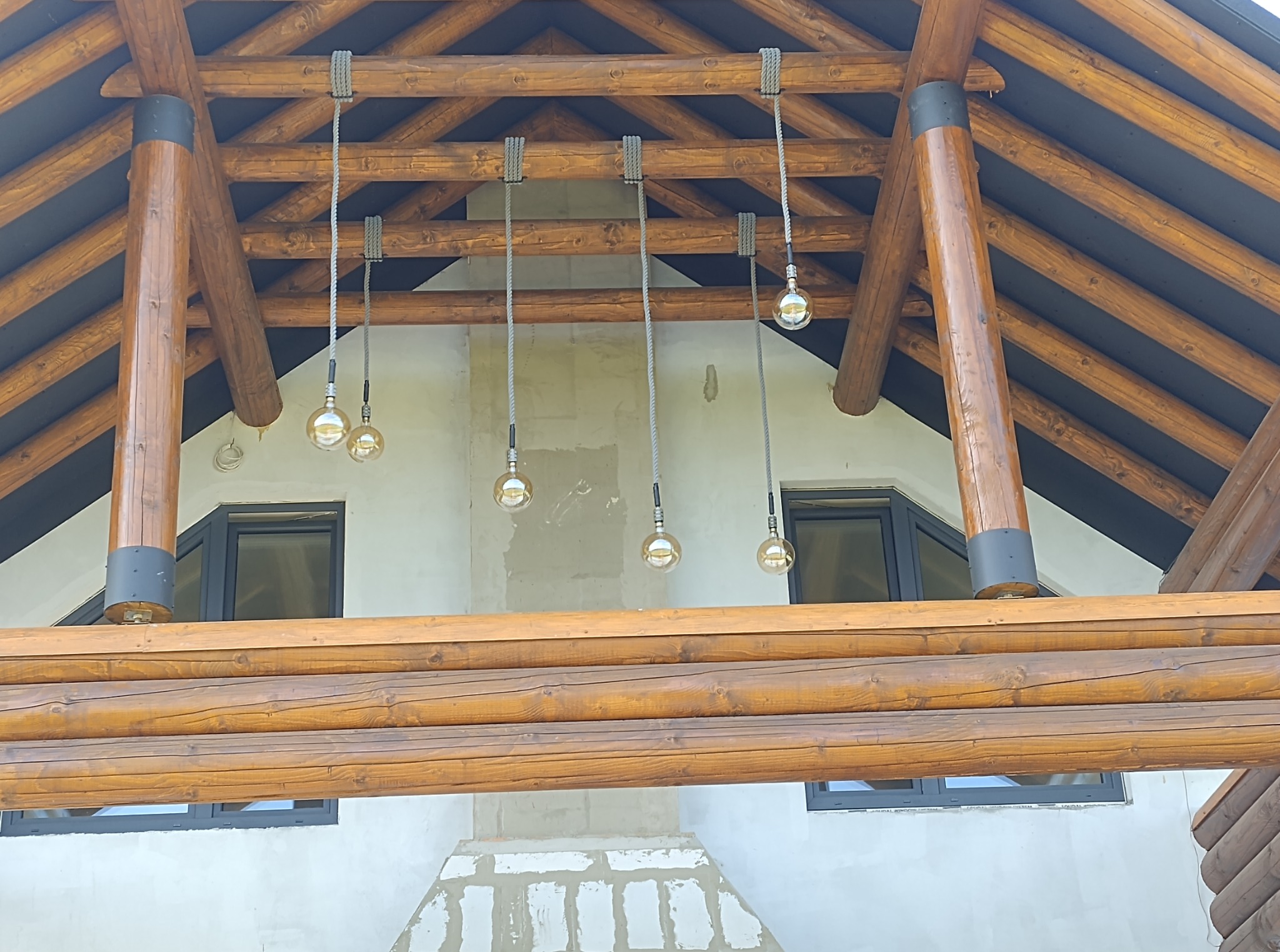 Drewniana konstrukcja dachu z oświetleniem w stylu retro. Lampy wiszące na sznurach, widoczne okna i fragment ściany budynku w trakcie wykończenia.