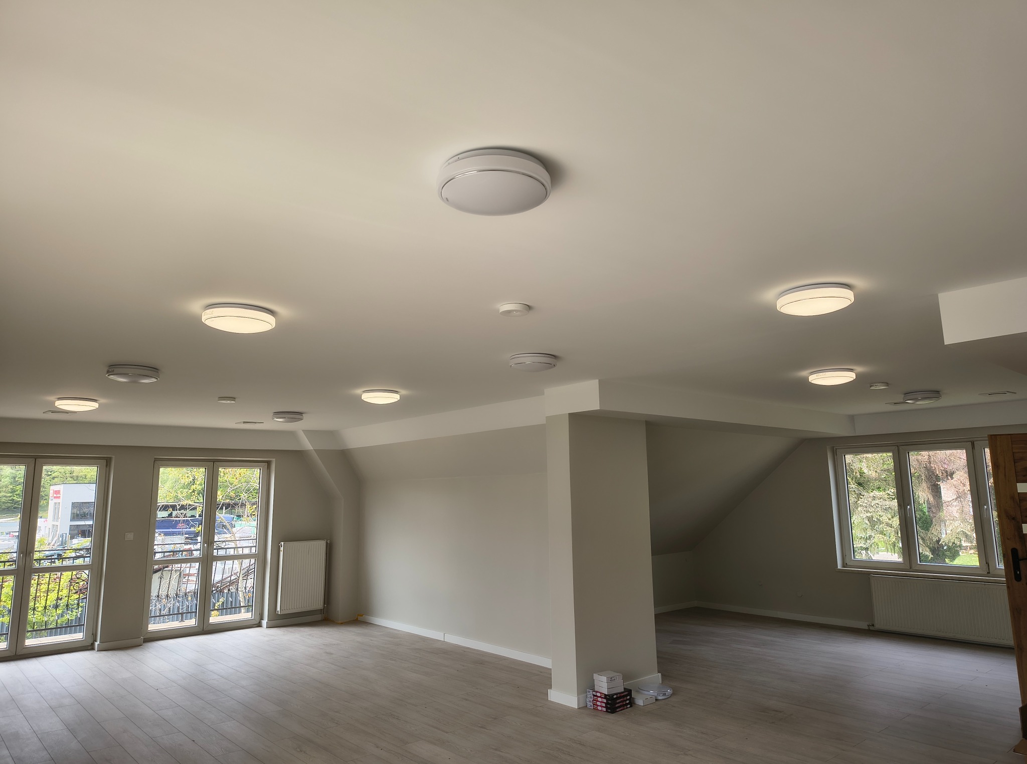 Jasne wnętrze poddasza z zamontowanymi okrągłymi lampami sufitowymi i czujnikiem dymu. Widoczne okna balkonowe z ozdobnymi balustradami i grzejnik.