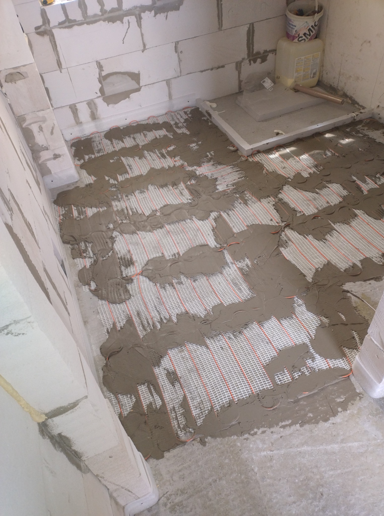 Układanie maty grzewczej na podłodze w surowym wnętrzu. Mata częściowo pokryta zaprawą, widoczne przewody grzewcze. Ściany z białych bloczków.