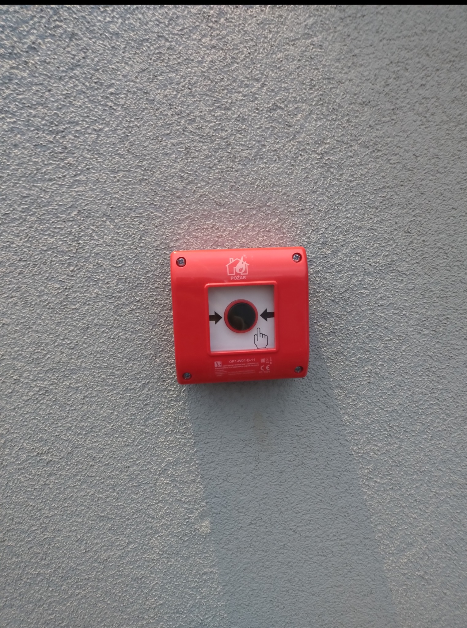 Czerwony, kwadratowy przycisk alarmowy 'POŻAR' zamontowany na szarej, teksturowanej ścianie. Widoczny symbol płomieni i dłoni wskazującej na czarny punkt.