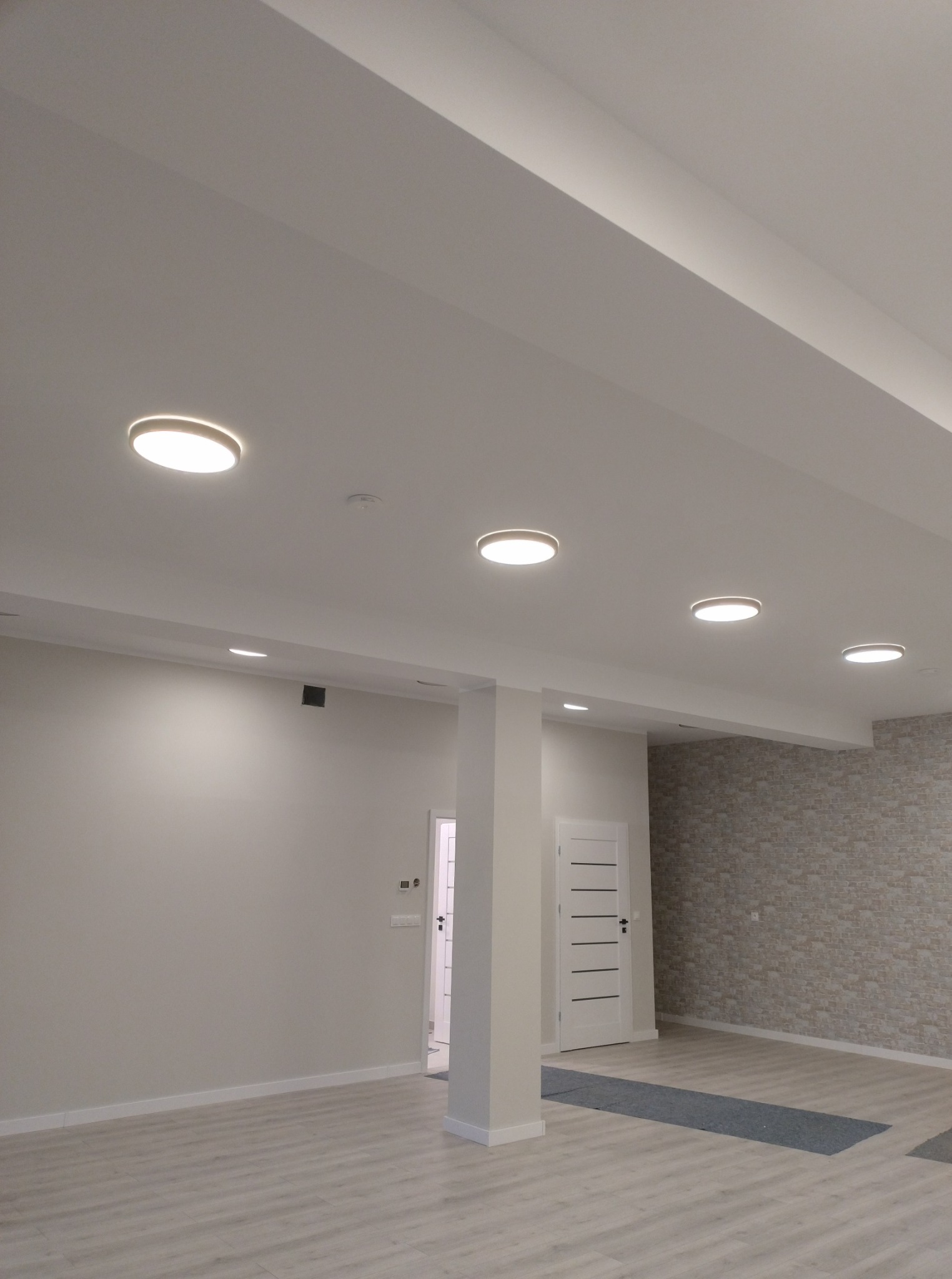 Wnętrze z zamontowanymi okrągłymi lampami sufitowymi LED, jasnymi ścianami i podłogą imitującą drewno. Widoczny element instalacji alarmowej na suficie.