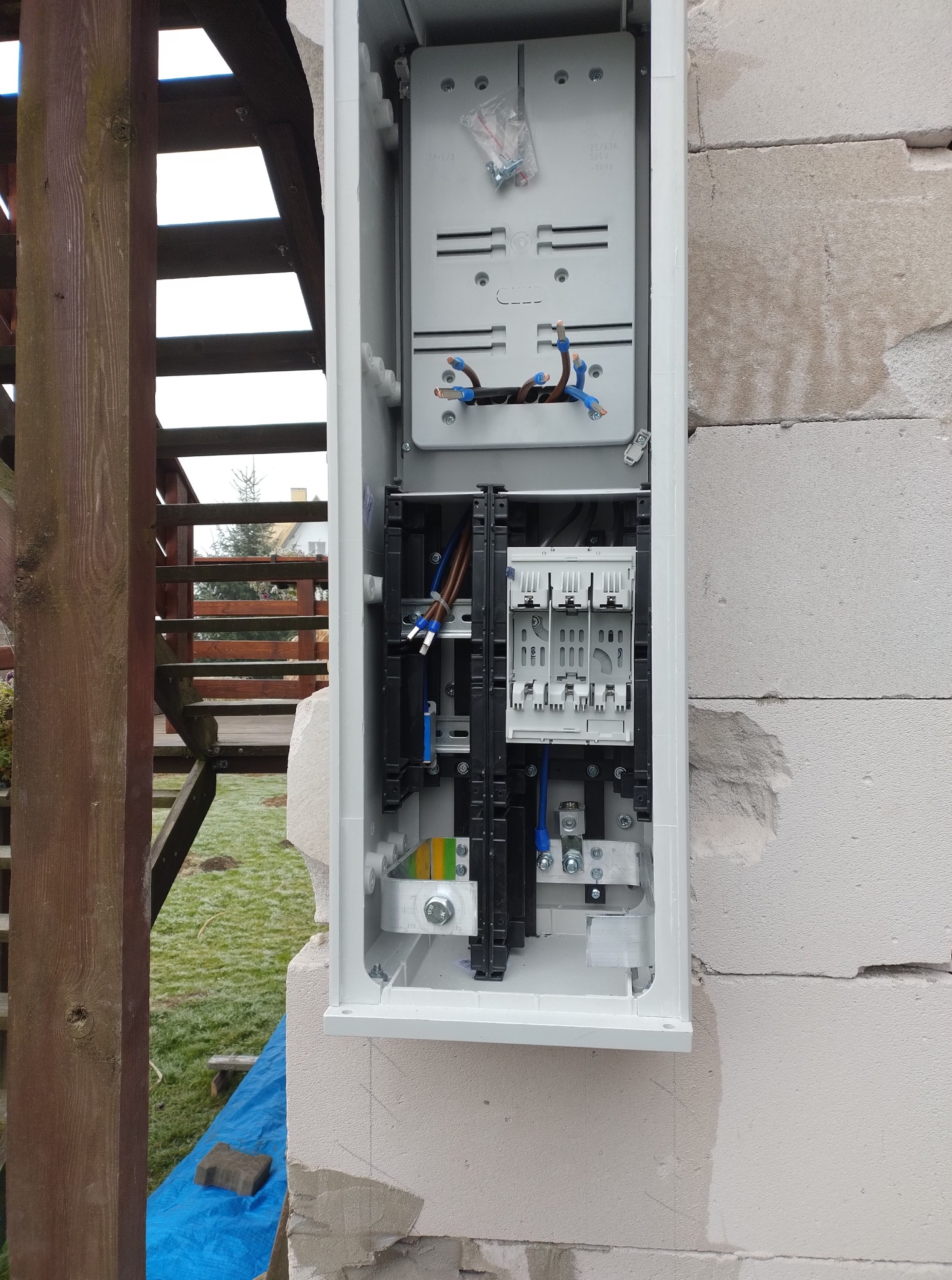 Szara skrzynka elektryczna zamontowana na ścianie z bloczków, widoczne kable i elementy instalacji. W tle drewniana konstrukcja i trawnik.