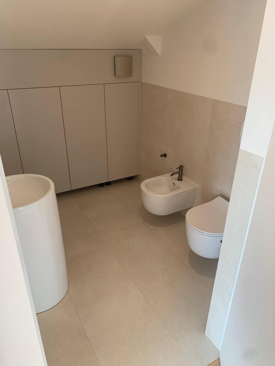 Minimalistyczna łazienka z białymi sanitariatami: umywalka kolumnowa, bidet i toaleta. Jasne płytki podłogowe i częściowo wykończone ściany. Proste, białe szafki w tle.