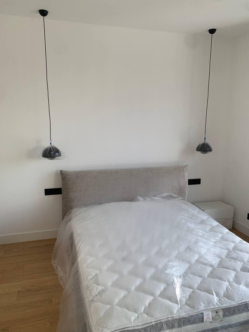 Pokój z łóżkiem zabezpieczonym folią i dwoma lampami wiszącymi z czarnymi kloszami, jeszcze w folii ochronnej. Białe ściany i drewniana podłoga. Przygotowanie do wykończenia.