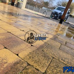 Czysty Ejber - Kostka brukowa po umyciu pod ciśnieniem, widoczne różnice w czystości. Część 'po' mokra i czysta, część 'przed' sucha i zabrudzona. W tle auto i zieleń.