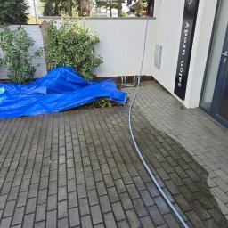 Czysty Ejber - Częściowo umyta kostka brukowa przed salonem urody, z wężem i niebieską plandeką. Widoczny efekt czyszczenia, odświeżony wygląd kostki.