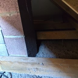 Krzysztof Cieślicki CK Izolacje - Poddasze z izolacją z wełny mineralnej, widoczne drewniane elementy konstrukcyjne i fragment komina. Izolacja wypełnia przestrzeń między krokwiami.