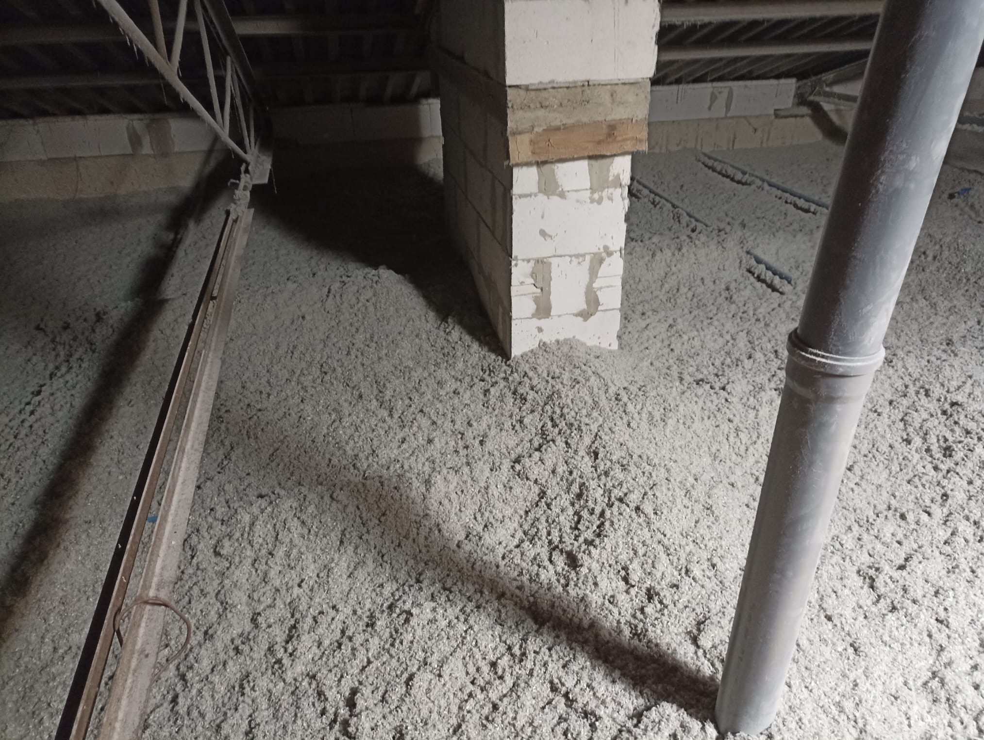 Poddasze z izolacją z wełny mineralnej, widoczny komin z bloczków i metalowa rura wentylacyjna. Ujęcie z dołu, widać konstrukcję dachu.