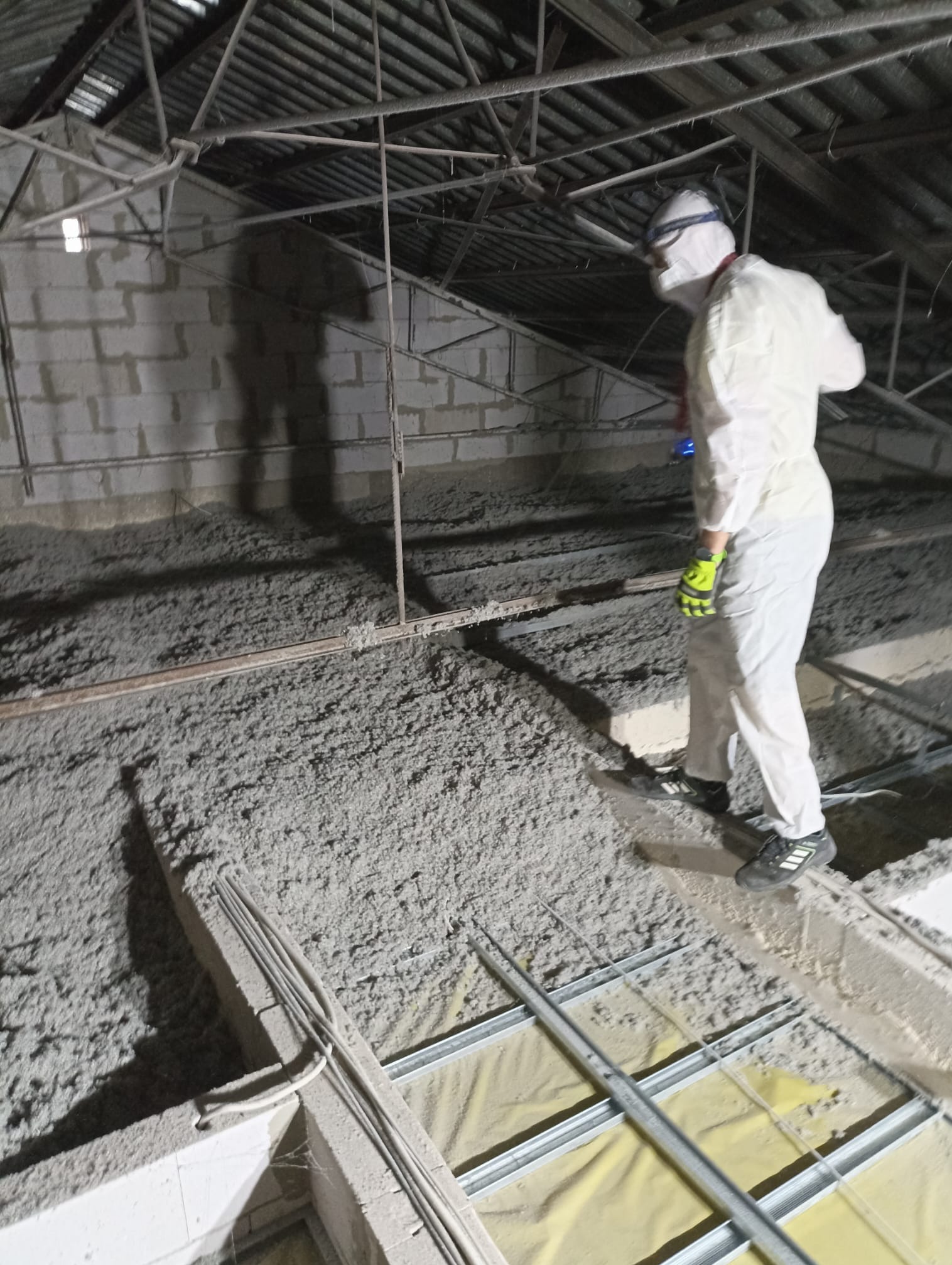 Pracownik w kombinezonie ochronnym ociepla poddasze. Widoczne belki konstrukcyjne i warstwa izolacyjna z wełny mineralnej. Oświetlenie robocze.