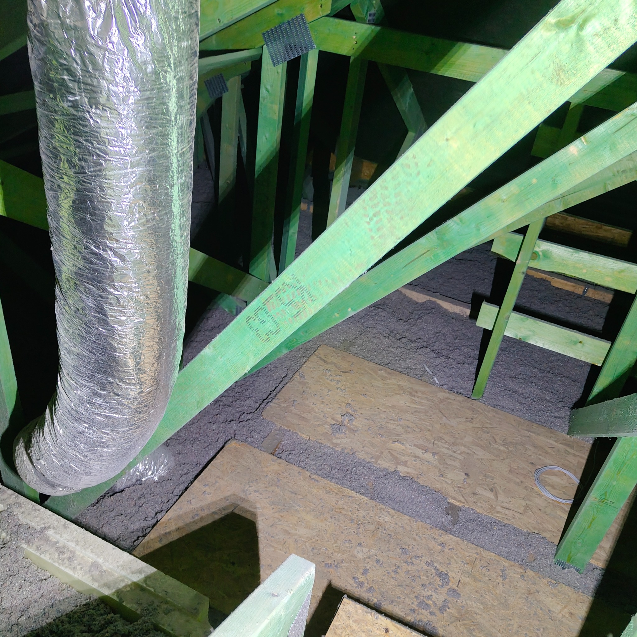 Poddasze z ociepleniem z wełny mineralnej, widoczne zielone elementy konstrukcji drewnianej i srebrny przewód wentylacyjny. Ocieplenie wykonane w przestrzeni między krokwiami.