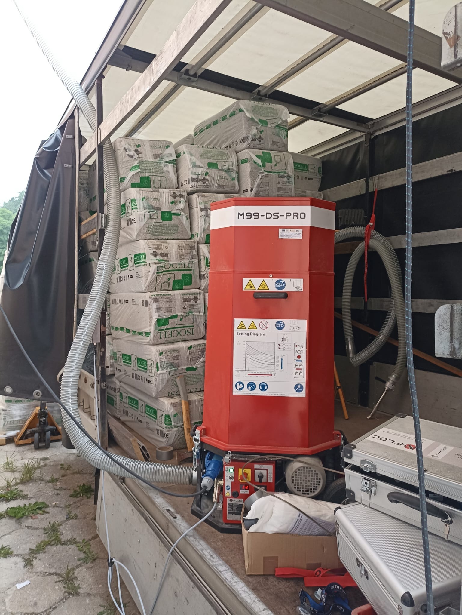 Maszyna M99-DS-PRO w przestrzeni ładunkowej, obok sterty worków z materiałem izolacyjnym Isocell. Widoczne węże i akcesoria, sugerujące przygotowanie do aplikacji.
