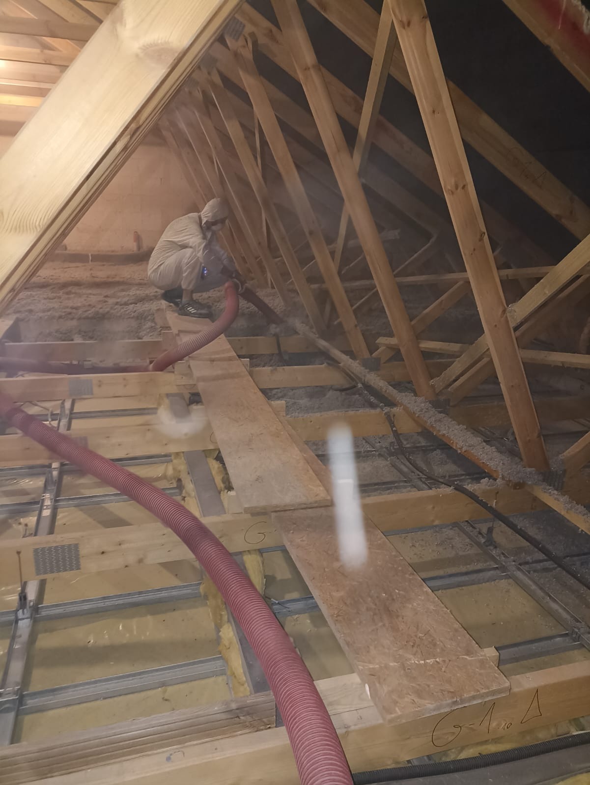 Pracownik w kombinezonie aplikuje izolację z czerwonego węża na drewnianą konstrukcję poddasza. Widoczne elementy konstrukcyjne i częściowo wypełniona przestrzeń izolacyjna.