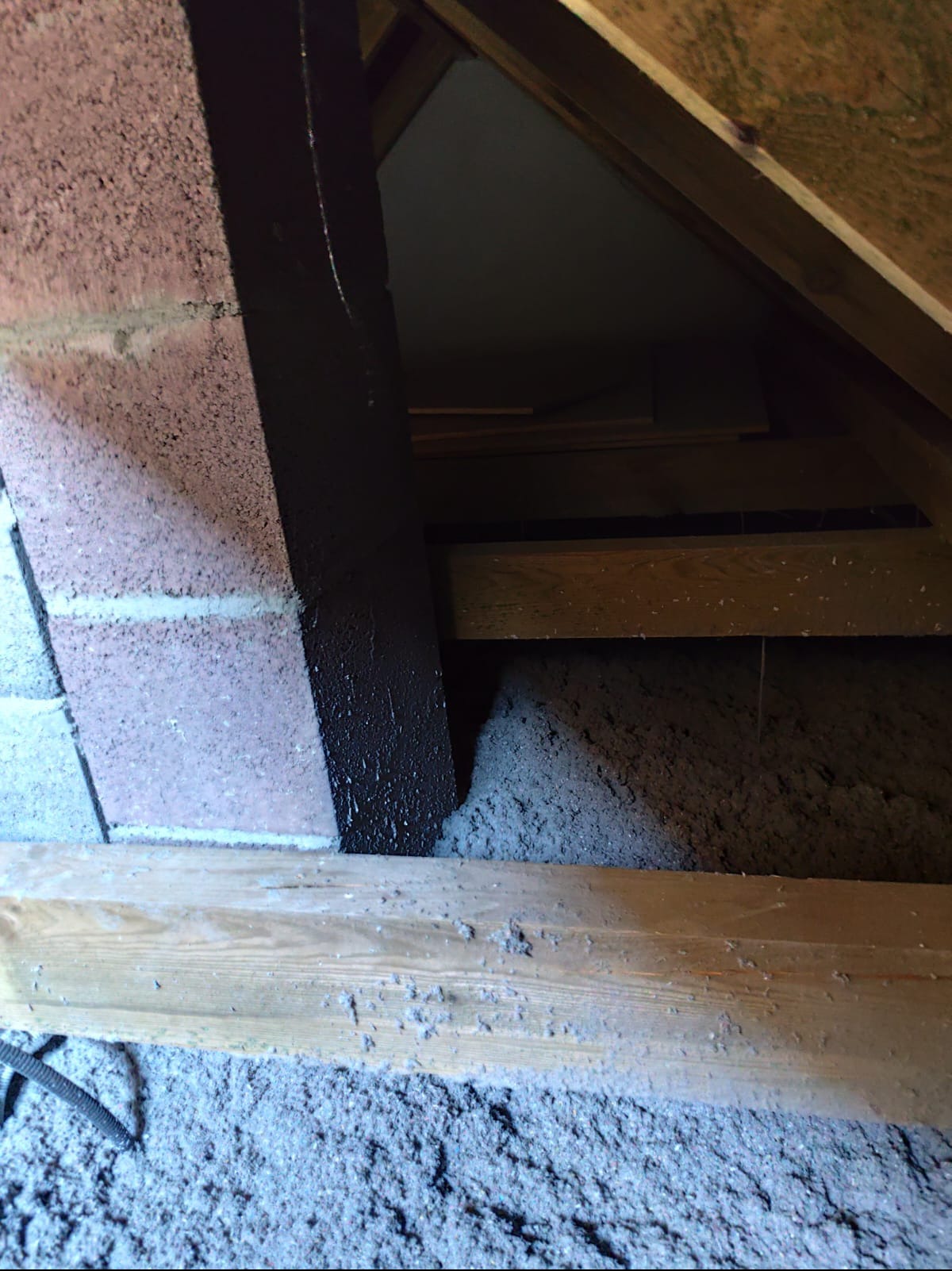 Poddasze z izolacją z wełny mineralnej, widoczne drewniane elementy konstrukcyjne i fragment komina. Izolacja wypełnia przestrzeń między krokwiami.