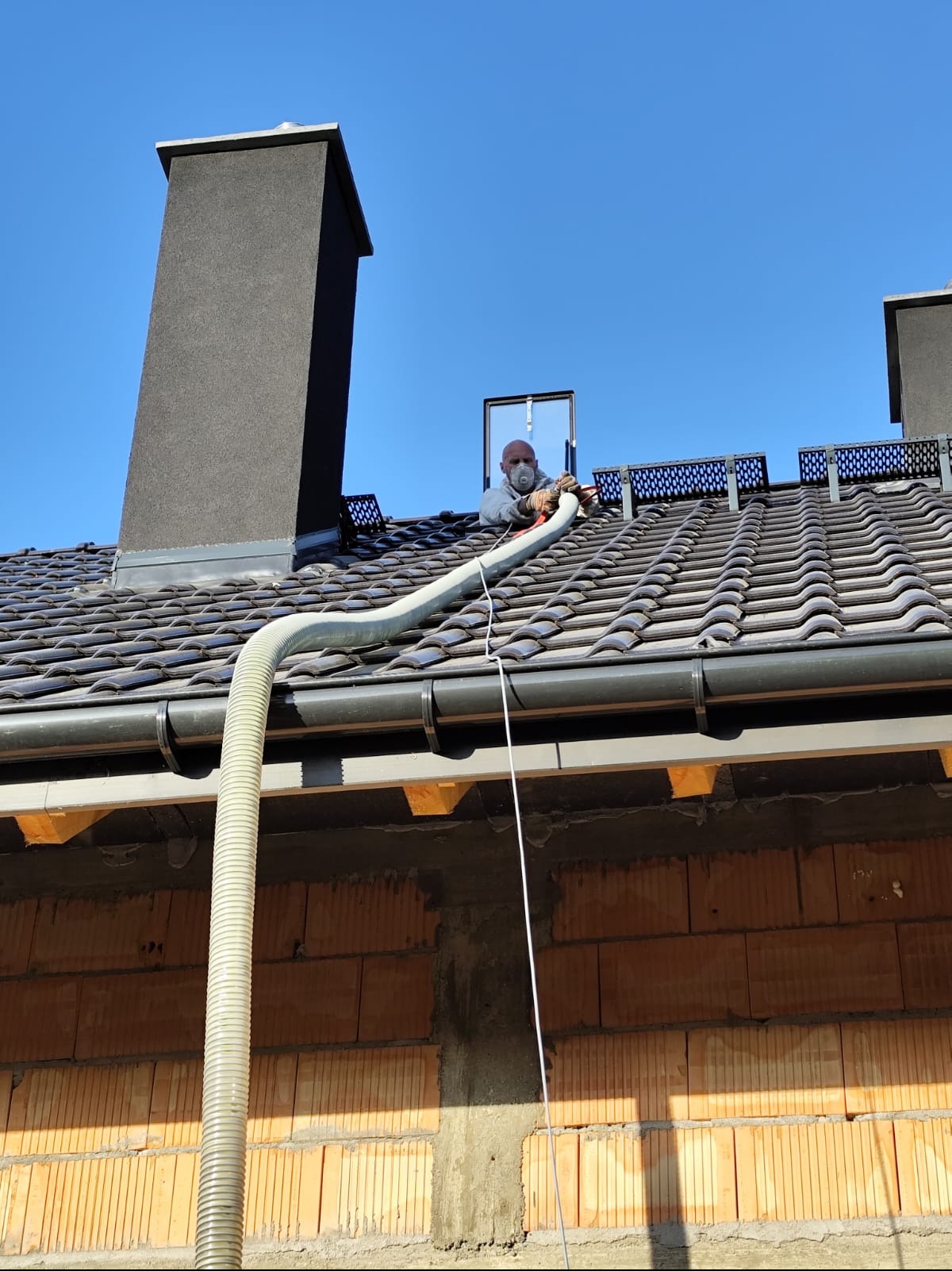 Pracownik w masce aplikuje izolację na poddaszu przez otwór w dachu. Widoczny komin, dachówka i fragment ściany z pustaków. Wąż do izolacji skierowany w stronę otworu.