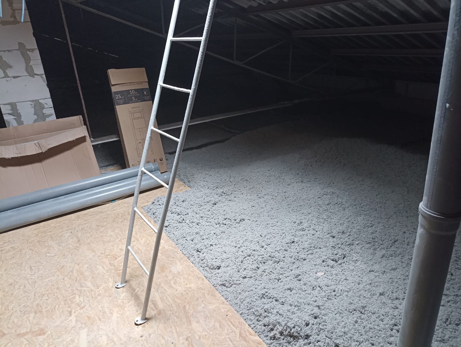 Poddasze z warstwą szarej izolacji, widoczna drabina, rury PCV i kartonowe pudła. Ujęcie z niskiego kąta, ukazujące strukturę materiału izolacyjnego.