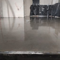 PAT-BET - Świeżo wylana, gładka posadzka przemysłowa w surowym wnętrzu. Widoczne instalacje na ścianach i zabezpieczenia folią. Błyszcząca powierzchnia odbija światło.