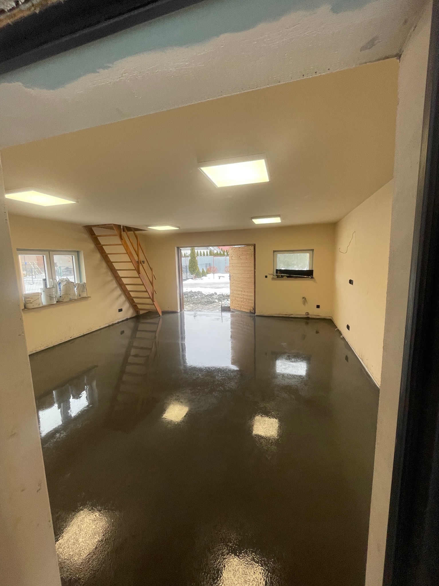 Błyszcząca, ciemna posadzka w jasnym pomieszczeniu z oknami i drewnianymi schodami. Widoczne worki z materiałami budowlanymi przy oknie. Otwarta brama garażowa.