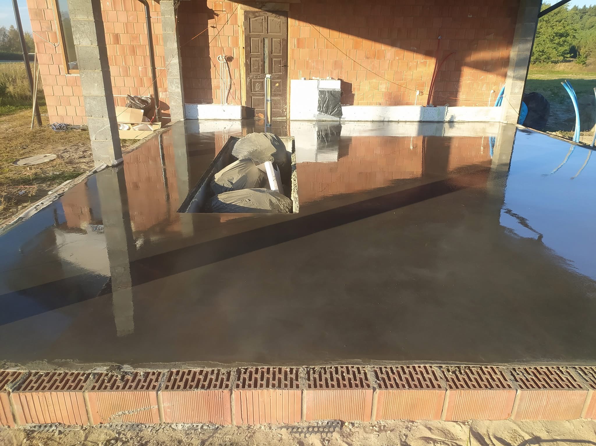 Świeżo wylana, gładka posadzka przemysłowa w surowym stanie, z widocznym odpływem liniowym i ceglanym obramowaniem. W tle fragment budynku w trakcie budowy.