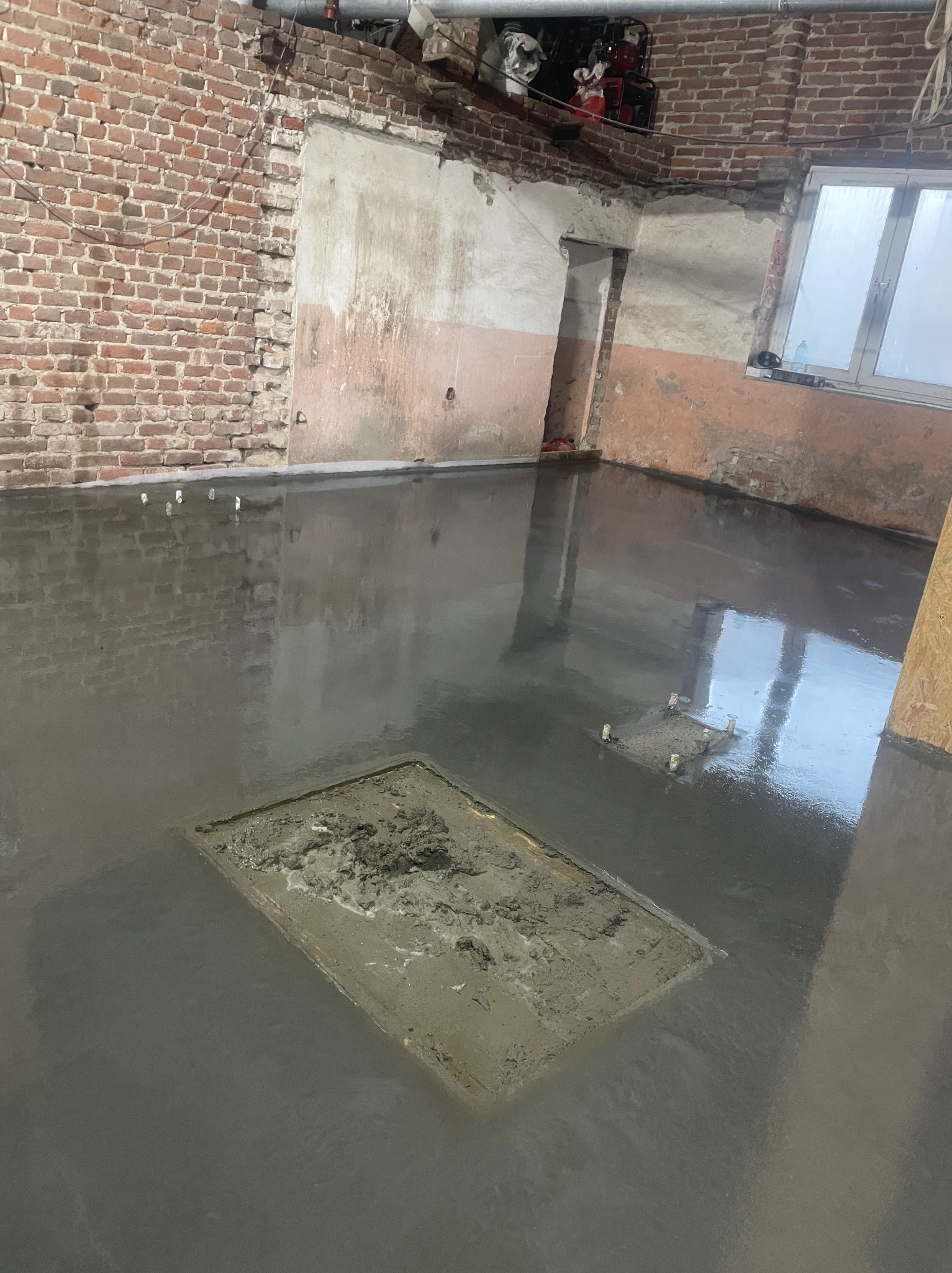 Świeżo wylana, gładka posadzka przemysłowa w surowym wnętrzu z ceglanymi ścianami. Widoczny właz rewizyjny w posadzce. Odbicie okna w mokrej powierzchni.