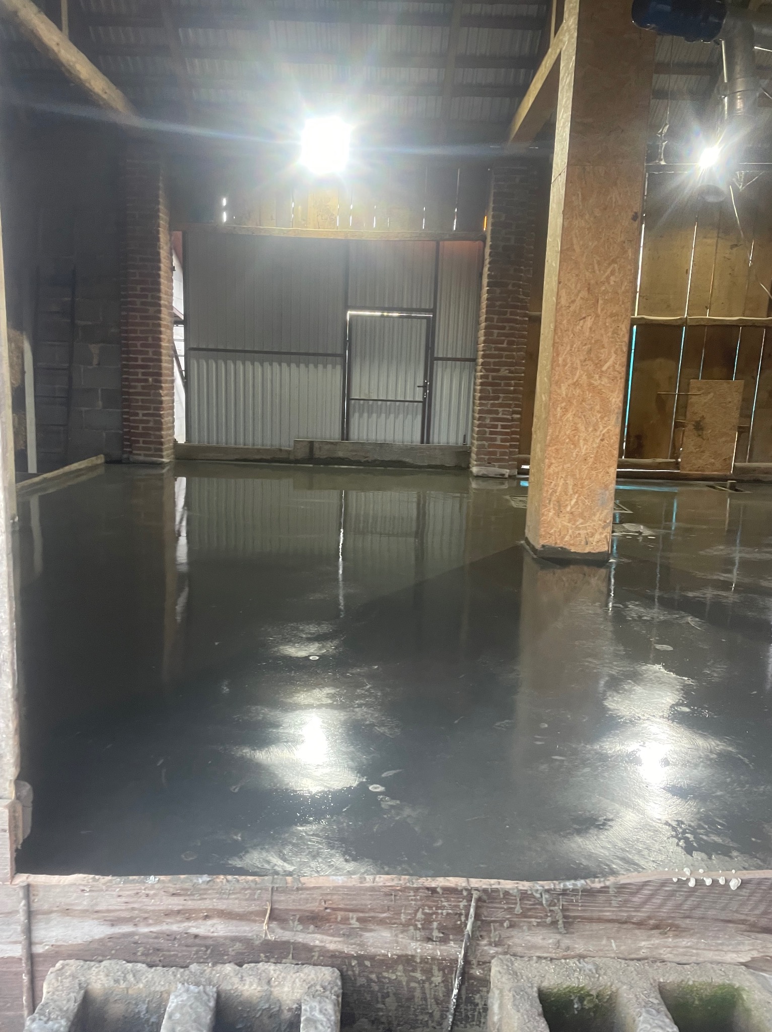 Wylana, mokra posadzka przemysłowa w surowym wnętrzu z ceglanymi filarami i drewnianą konstrukcją. Widoczne odbicia światła na gładkiej powierzchni.