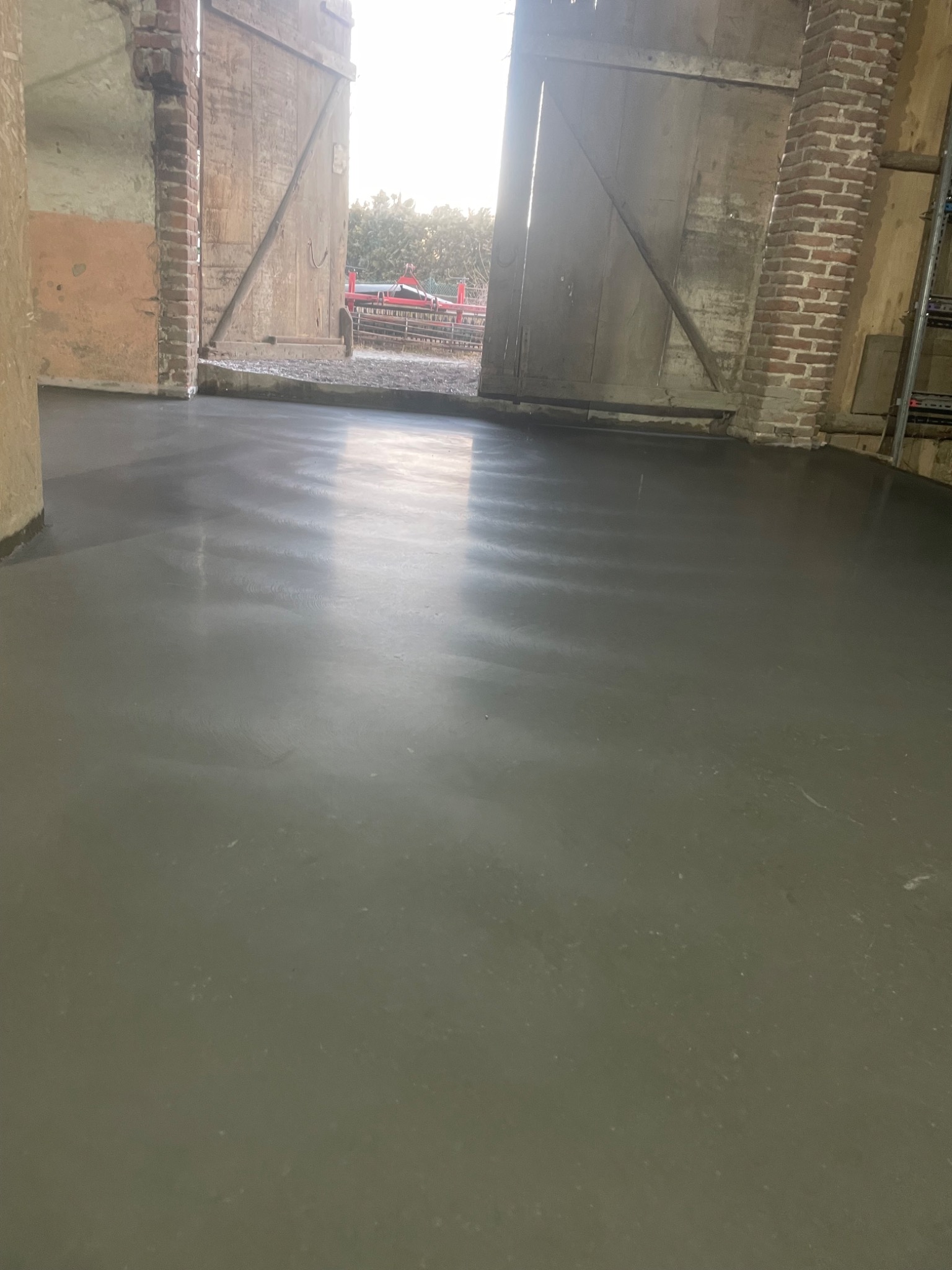 Gładka, szara posadzka przemysłowa wewnątrz budynku gospodarczego z ceglanymi filarami i otwartymi drewnianymi wrotami, przez które widać zieleń i czerwony sprzęt rolniczy.