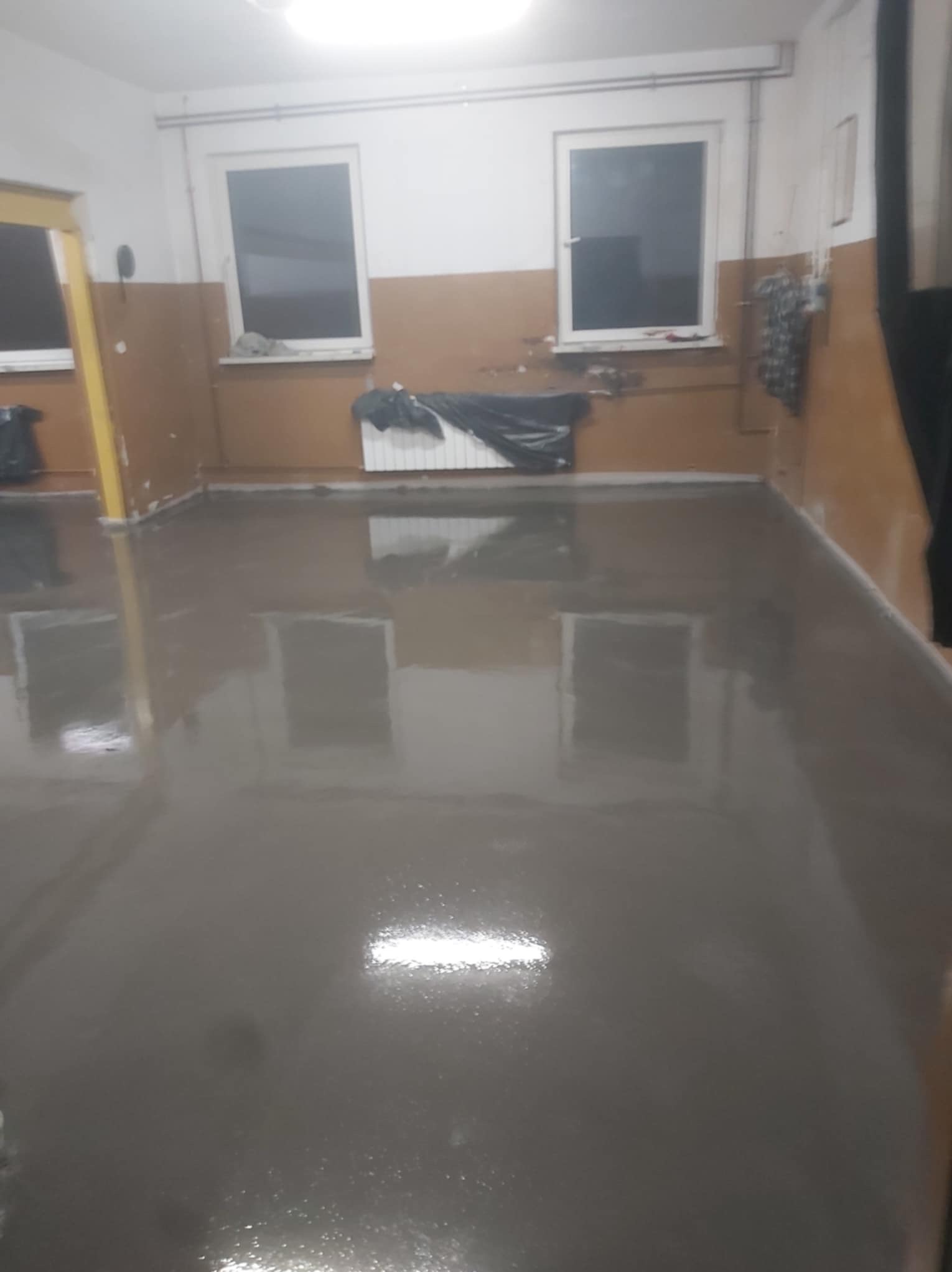 Świeżo wylana, gładka, szara posadzka w pomieszczeniu z dwoma oknami. Na parapecie widoczne zabezpieczenia. Połyskująca powierzchnia odbija światło.