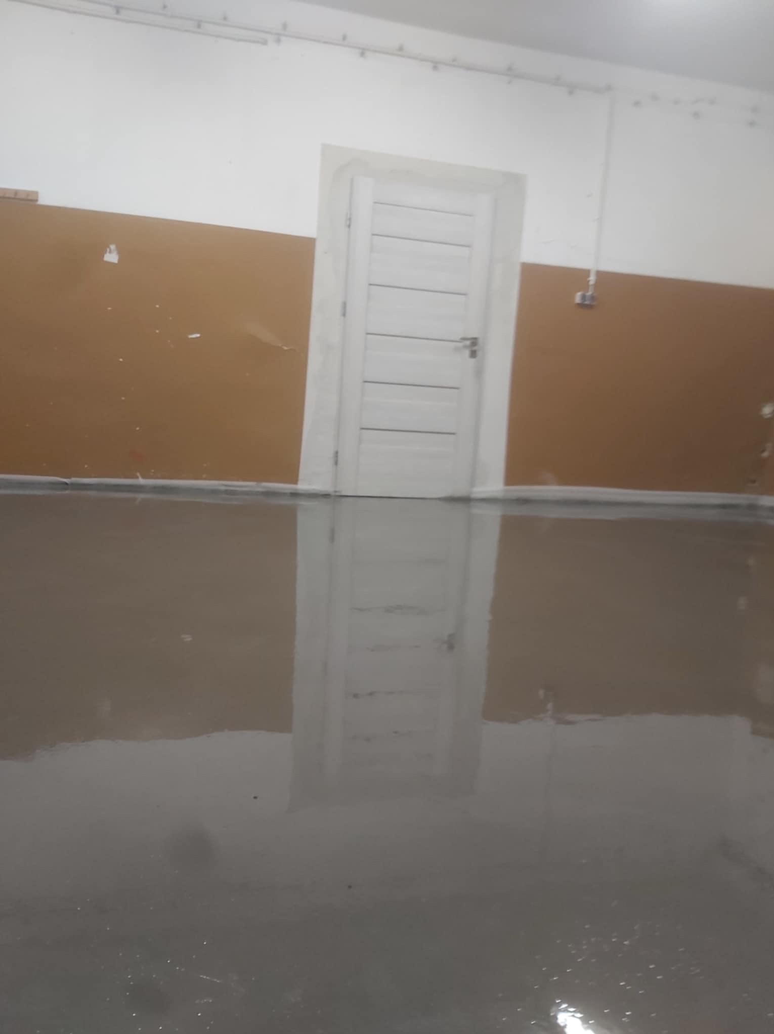 Świeżo wylana, gładka, szara posadzka samopoziomująca w pomieszczeniu z biało-brązowymi ścianami i białą, drewnianą ościeżnicą drzwiową.