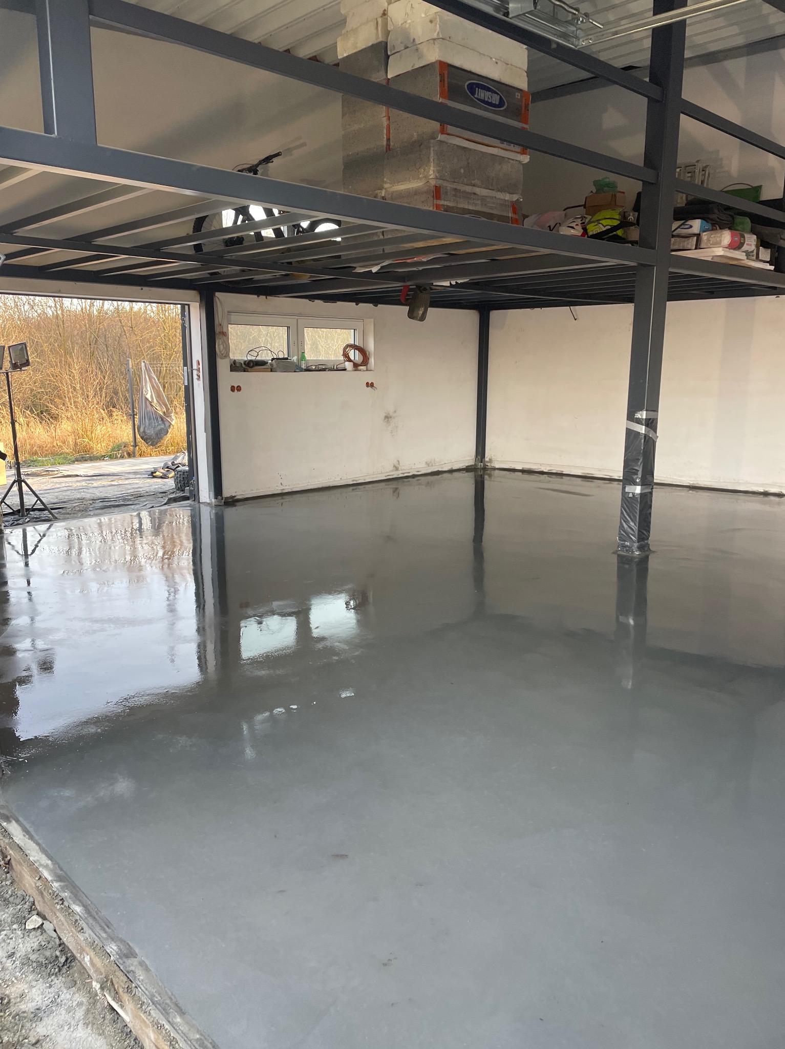 Świeżo wylana, gładka, szara posadzka w surowym wnętrzu z antresolą. Widoczne refleksy światła na powierzchni, surowe ściany i stalowa konstrukcja antresoli.